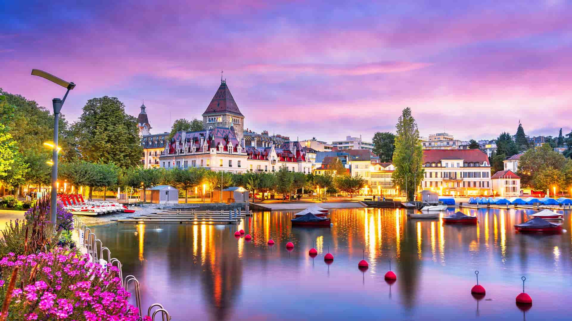"A breathtaking sunset shot of the waterfront in Lausanne, Switzerland, with the city's charming architecture reflecting in the still waters of Lake Geneva, framed by pink flowers and moored boats.  "