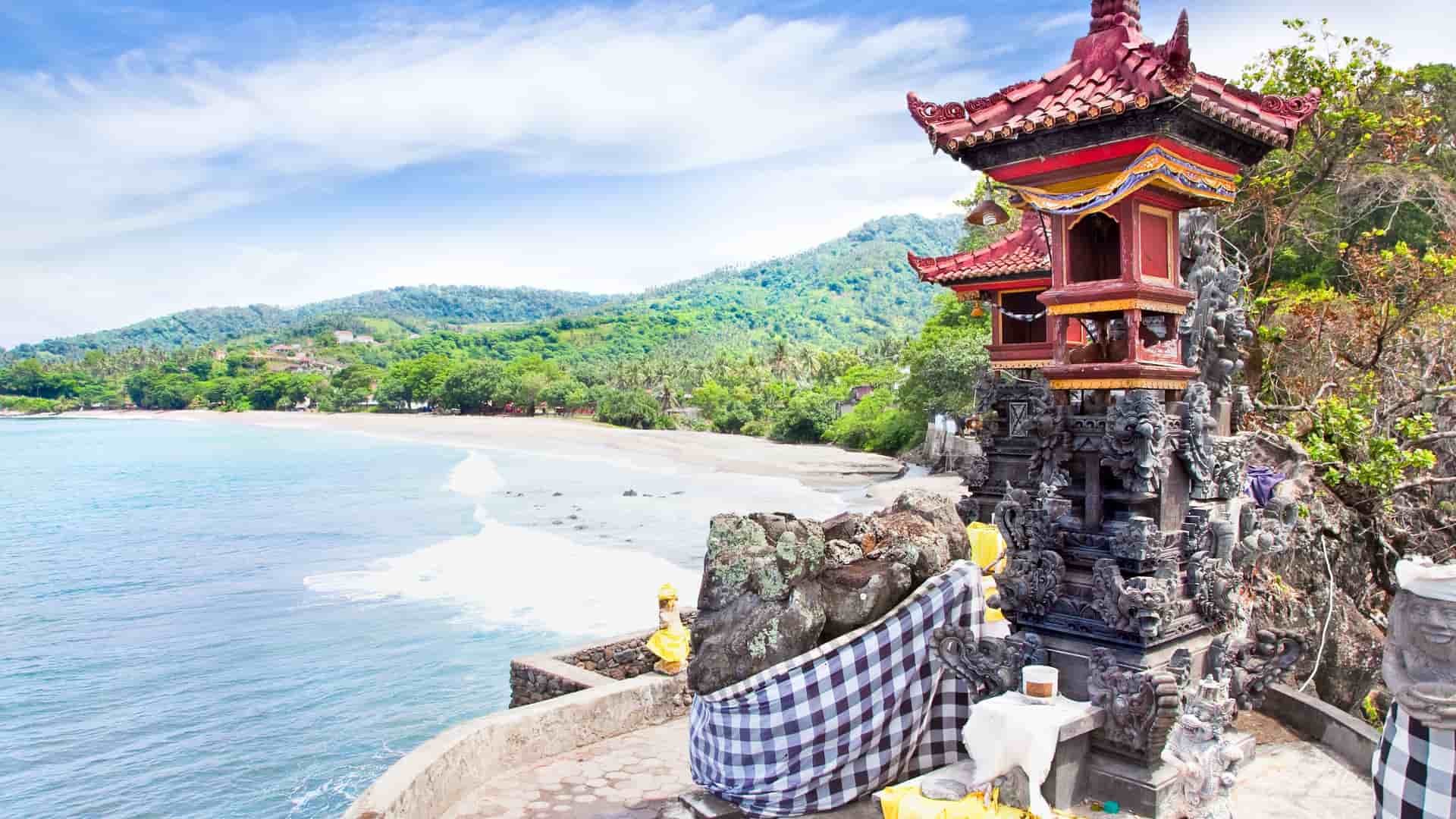 A wide shot of a traditional Balinese temple on a cliff overlooking a beautiful beach with white sand and turquoise water on the tropical island of Lombok, Indonesia.