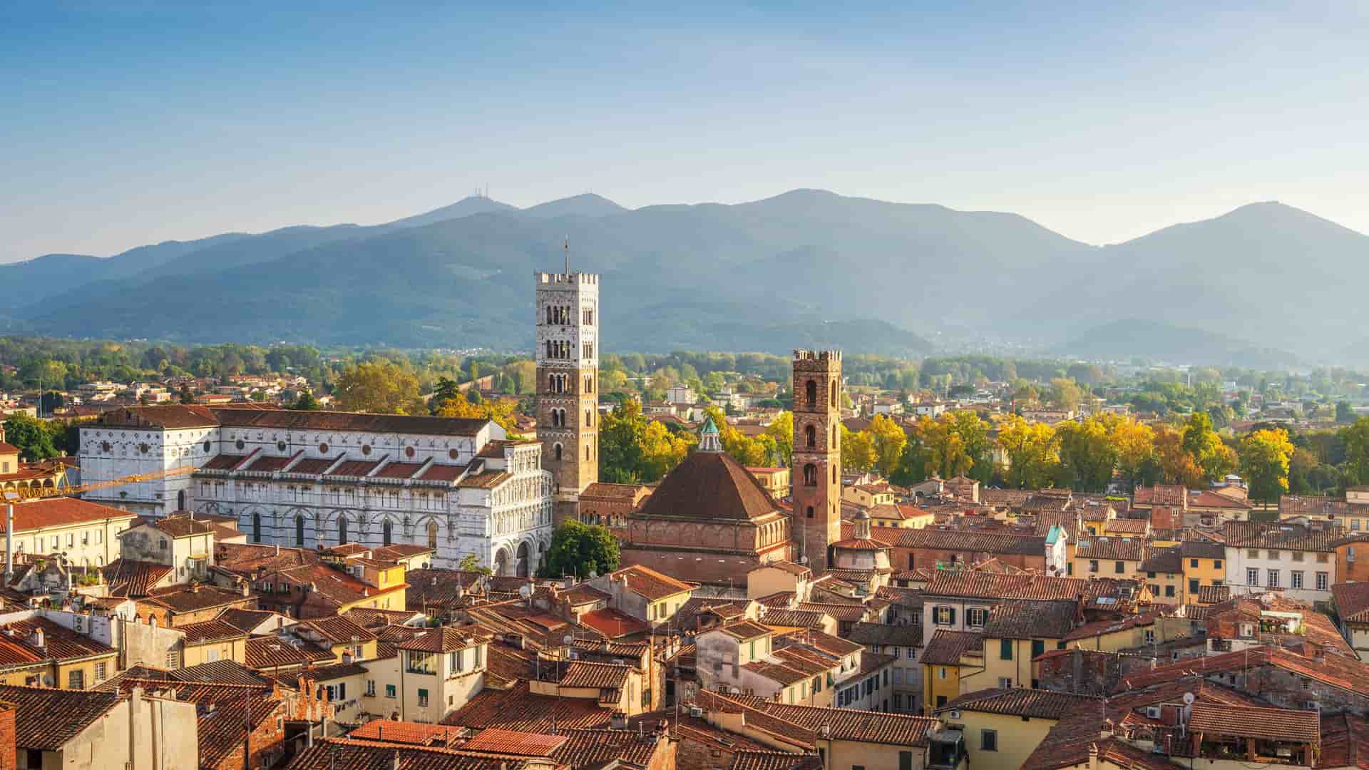 "An aerial view of the historic city of Lucca, Italy, with its terracotta rooftops, prominent bell towers, and a backdrop of rolling Tuscan hills.  "