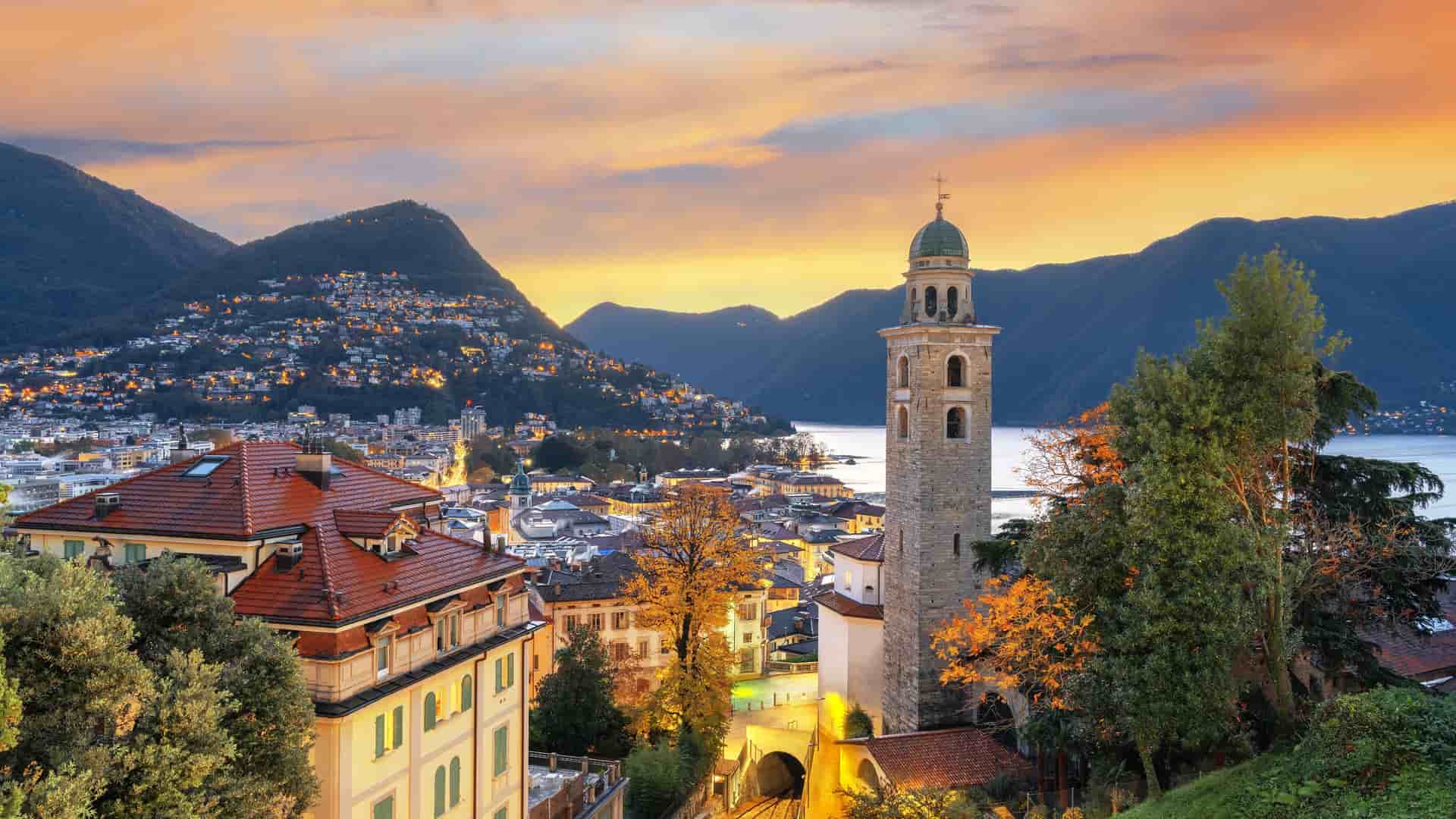 "An elevated sunset view of Lugano, Switzerland, highlighting the beautiful bell tower of the Church of Santa Maria degli Angeli, with Lake Lugano and the surrounding mountains in the background.  "