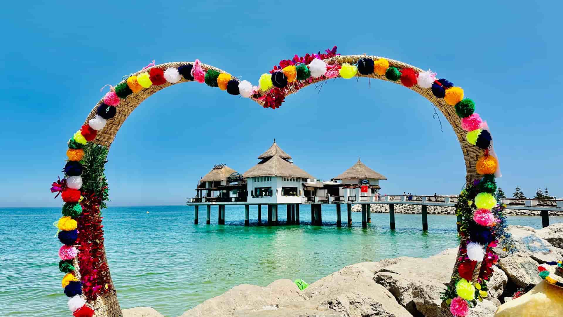 "A large heart-shaped arch decorated with colorful pom-poms frames a view of a tranquil blue sea with a stilted overwater restaurant in M'diq, Morocco. "