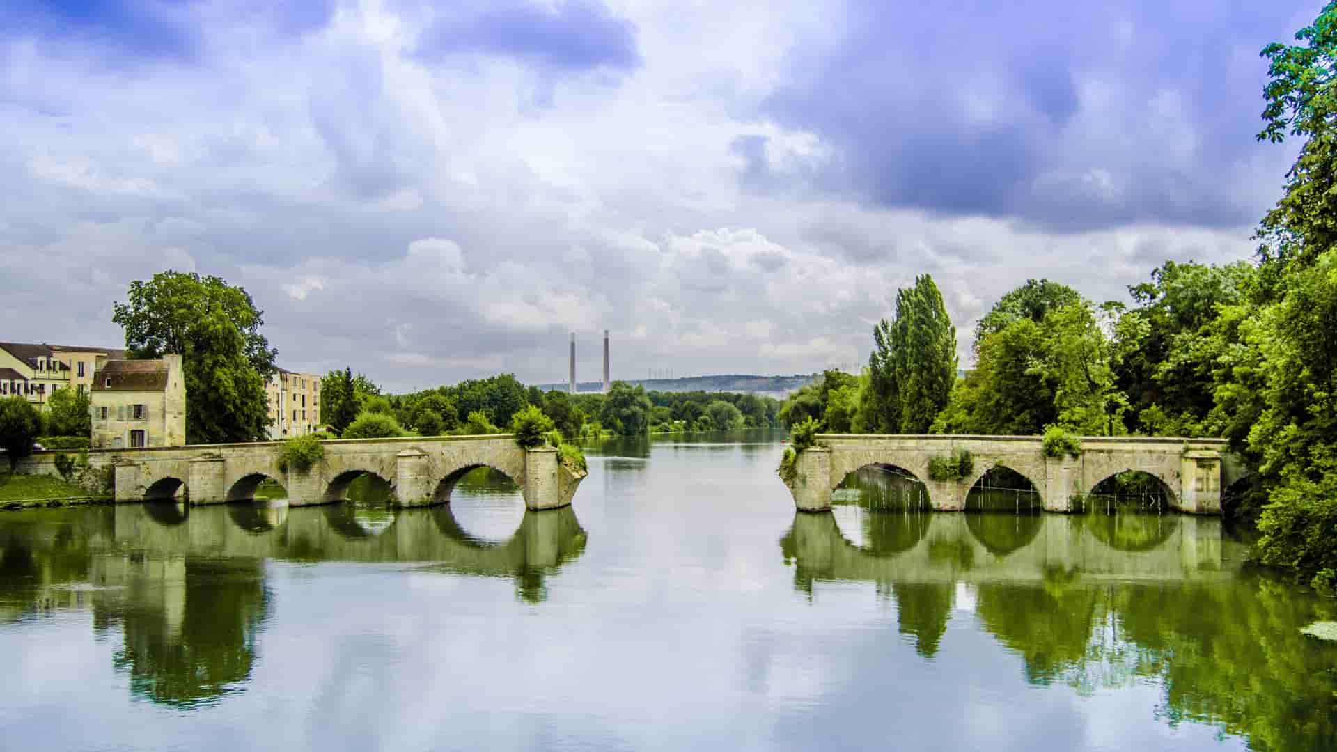 A picturesque view of the old stone bridge with multiple arches spanning the calm waters of the Seine River in Mantes-la-Jolie, France.