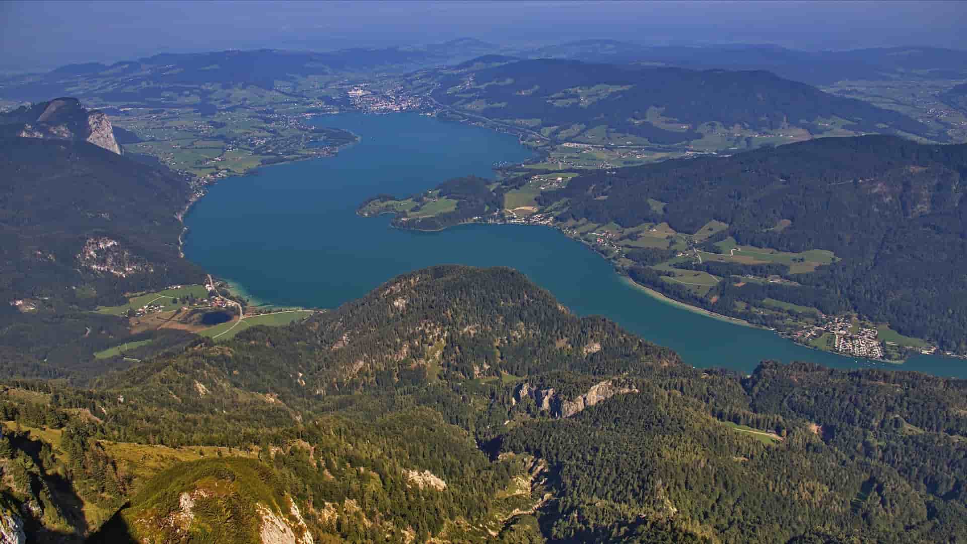 An aerial view of the serene, crescent-shaped Mondsee lake in Austria, surrounded by the lush green valleys and towering, tree-covered mountains of the Salzkammergut region.