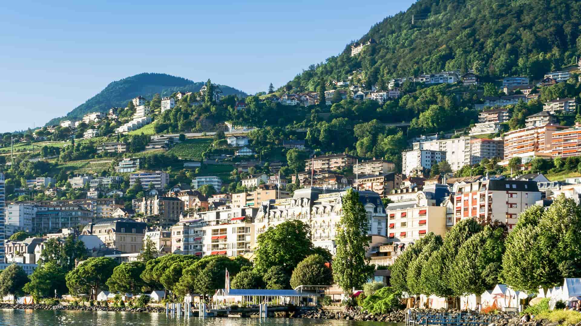 A panoramic view of Montreux, Switzerland, with colorful buildings lining the shores of Lake Geneva and climbing the steep, green hillsides behind the city.