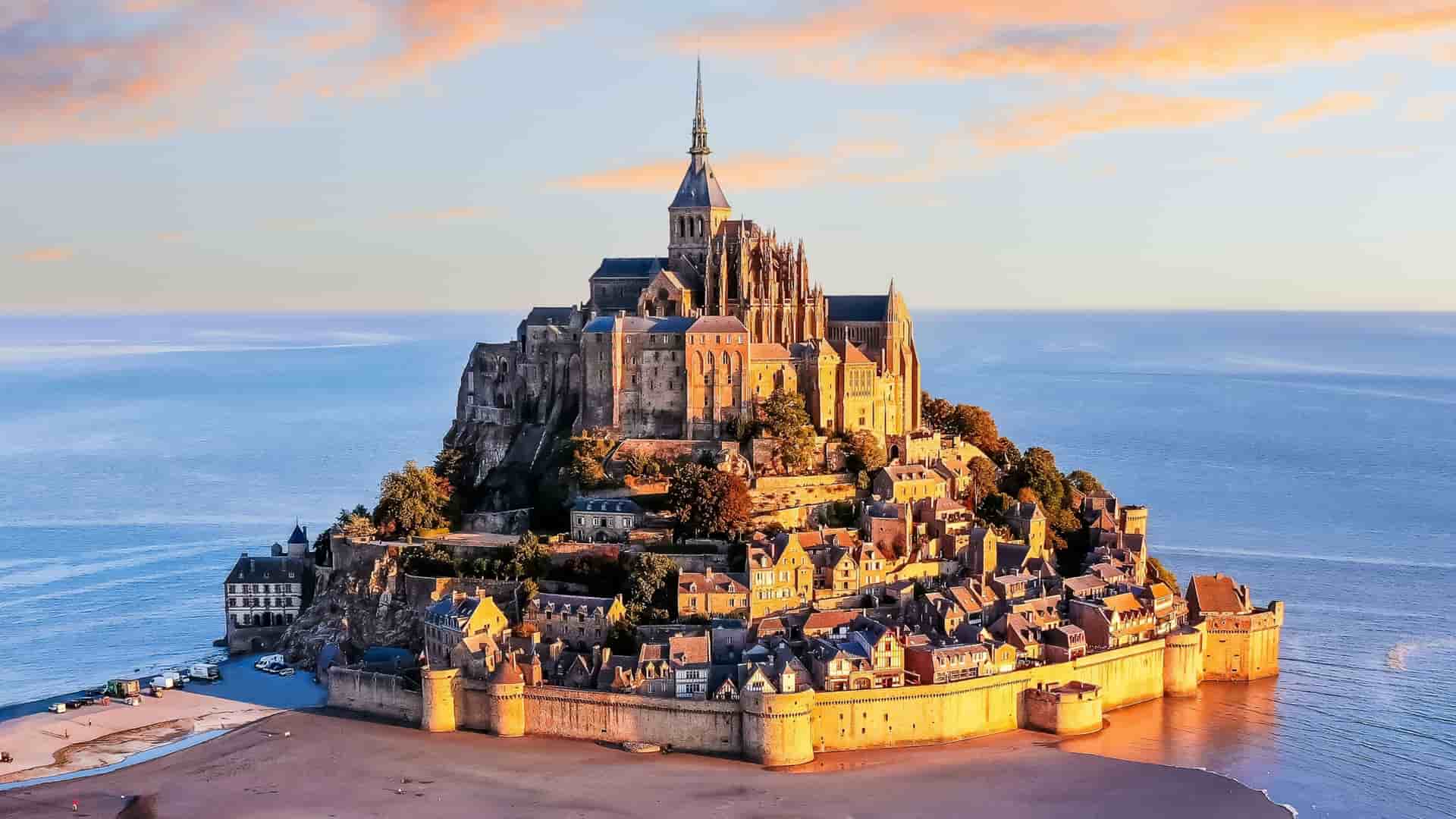 A high-angle view of the iconic Mont Saint-Michel, a tidal island and commune in Normandy, France, with its stunning medieval abbey and village rising from the sea at sunset.