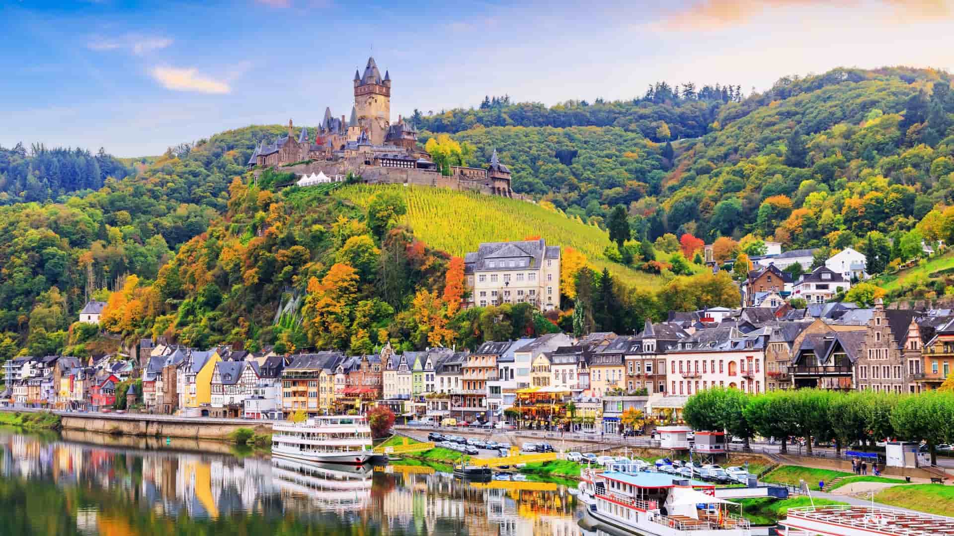 A picturesque view of Cochem, Germany, with the historic Reichsburg Castle perched on a hill above the colorful town and the Moselle River, with cruise ships docked along the embankment.