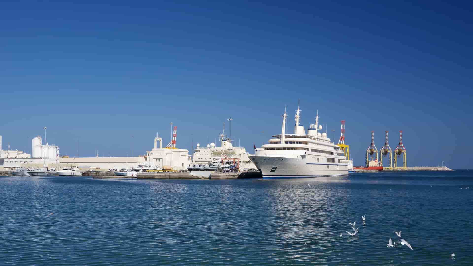 A high-angle view of Port Sultan Qaboos in Muscat, Oman, with a large cruise ship or cargo vessel docked in the harbor. The port is backed by the residential buildings of the city's waterfront, which climb the surrounding rugged, arid mountains. The scene captures the commercial and scenic beauty of Muscat's main port.