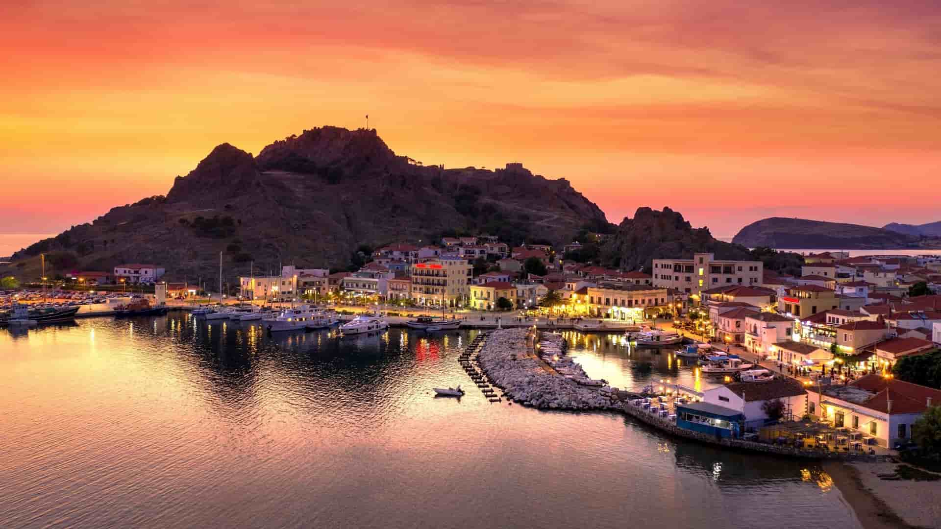 A vibrant sunset view of the harbor and town of Myrina, Greece, with boats docked and lights illuminating the buildings against a backdrop of a mountain with a castle ruin.