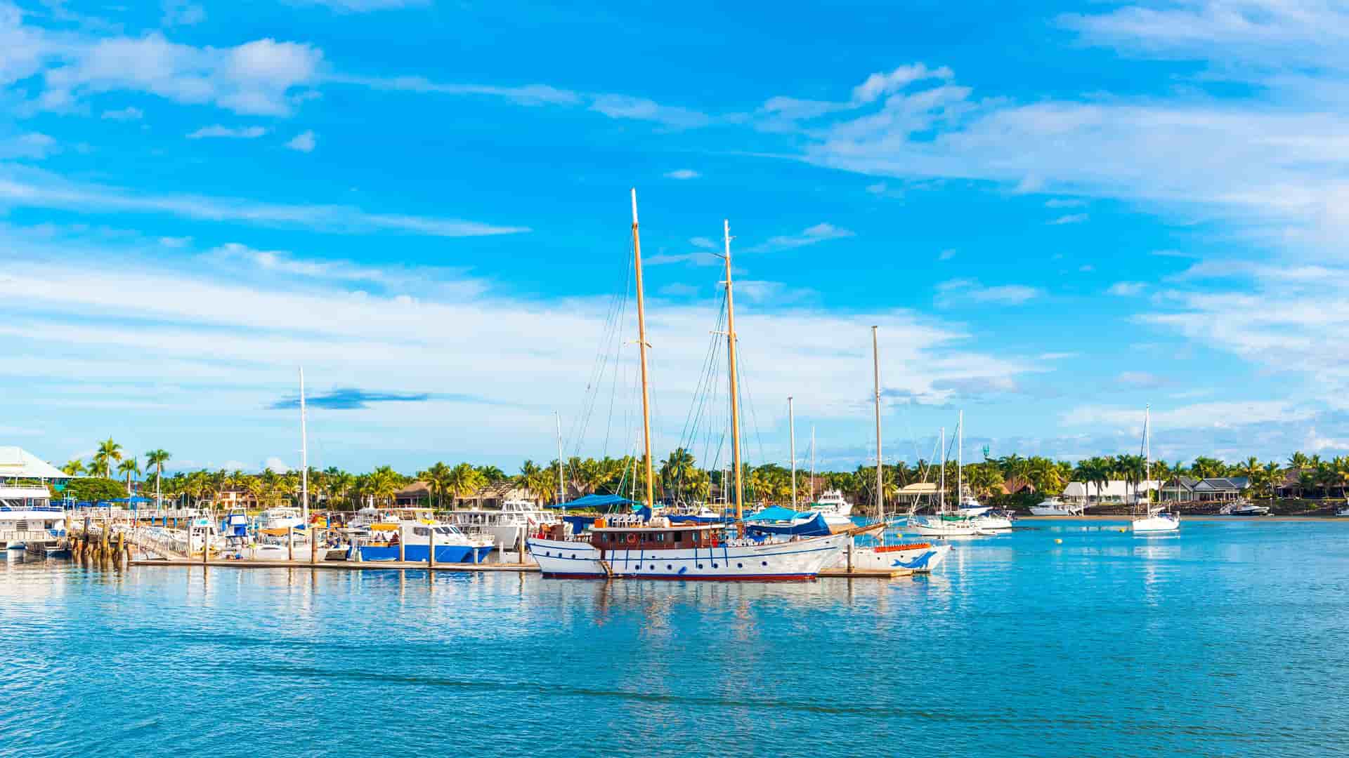 "A sunny, wide-angle shot of a marina in Nadi, Fiji, with several boats docked in the calm turquoise water under a bright blue sky with scattered clouds.  "