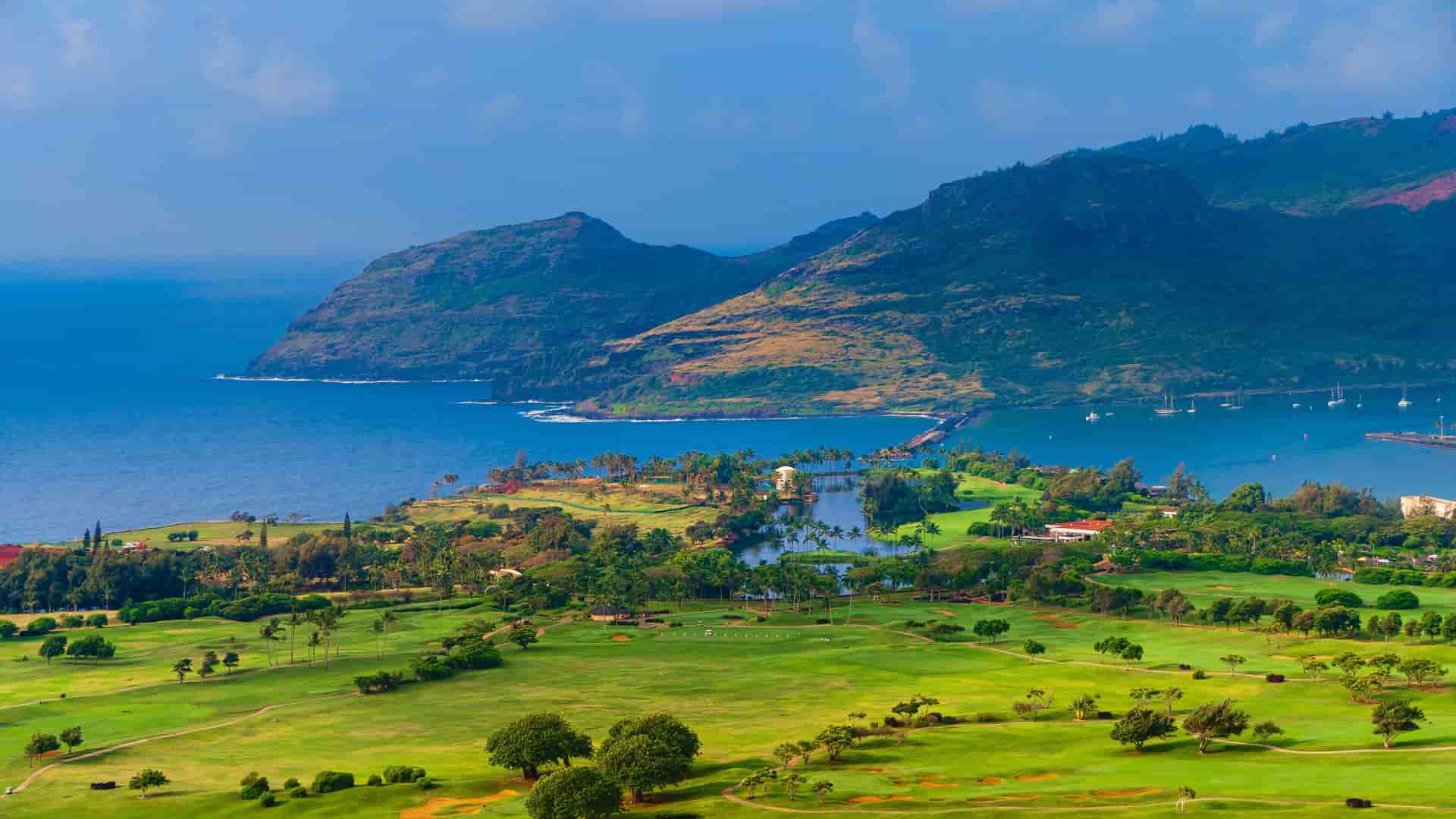 A panoramic view of a lush green golf course and resort area near Nawiliwili Harbor in Kauai, Hawaii, with the ocean and volcanic mountains in the background.