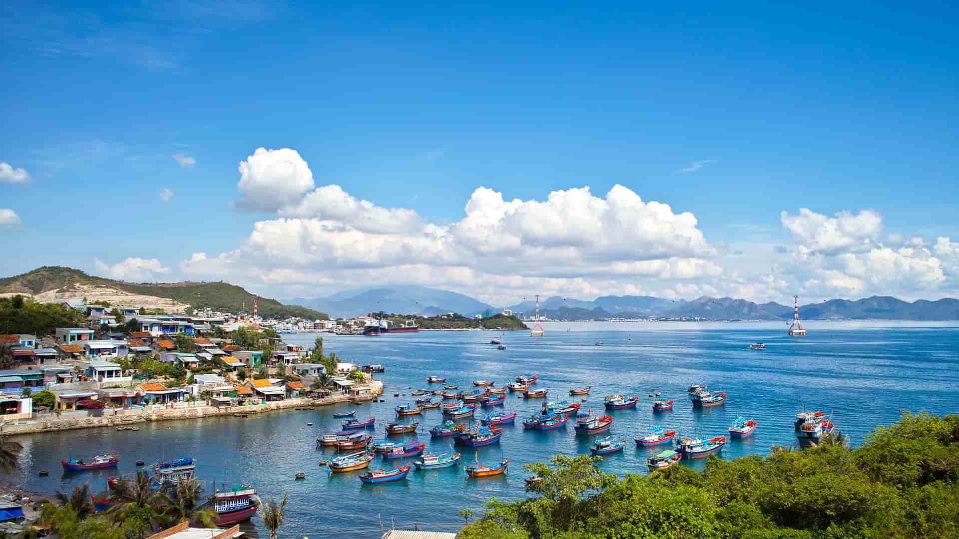 A panoramic view of Nha Trang, Vietnam, with a bustling fishing harbor full of colorful boats, a coastal town, and mountains in the distance under a bright blue sky.