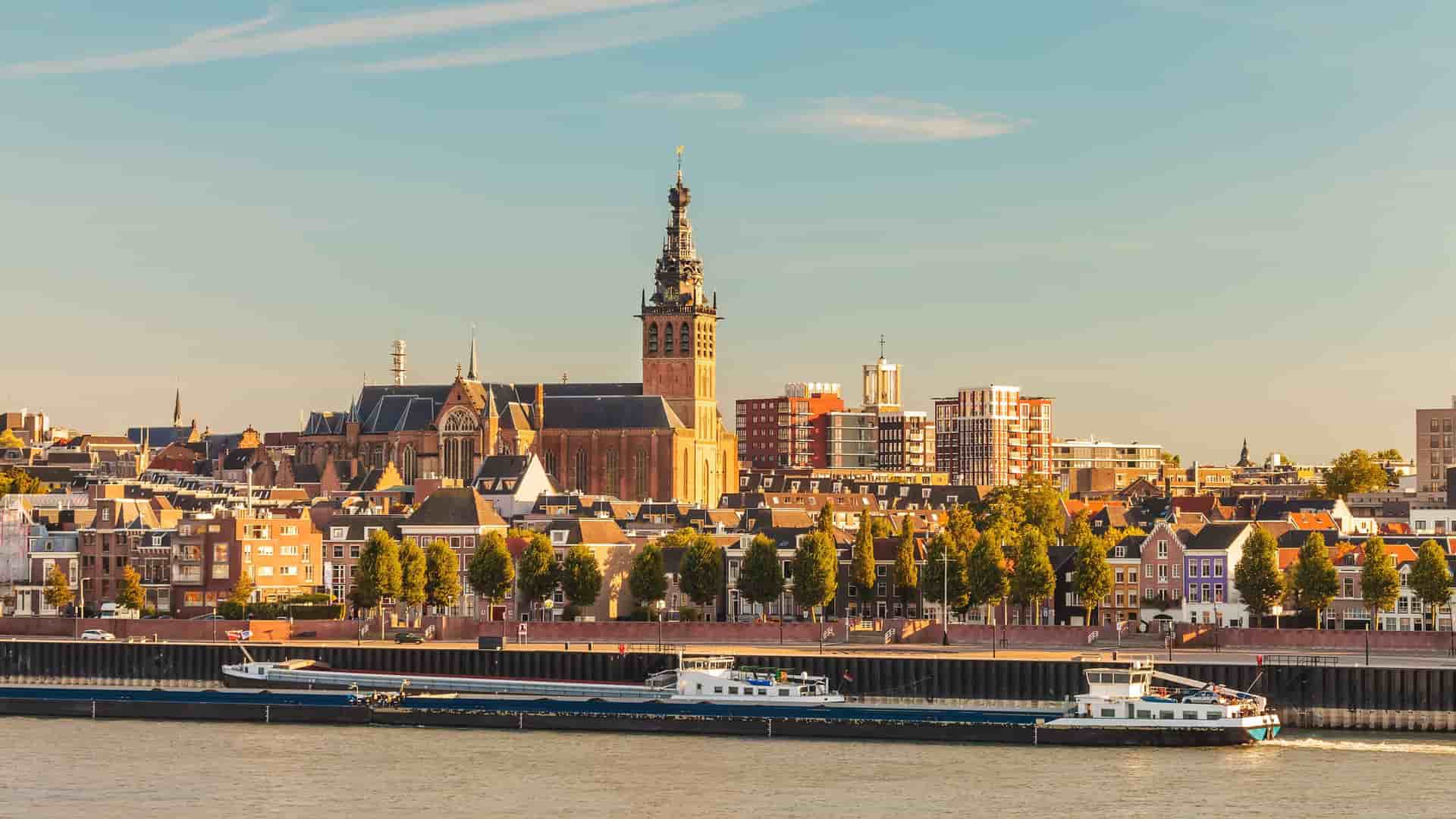 A river cruise barge navigates the Waal River with a panoramic view of the Nijmegen, The Netherlands skyline and the historic Stevenskerk church dominating the cityscape.