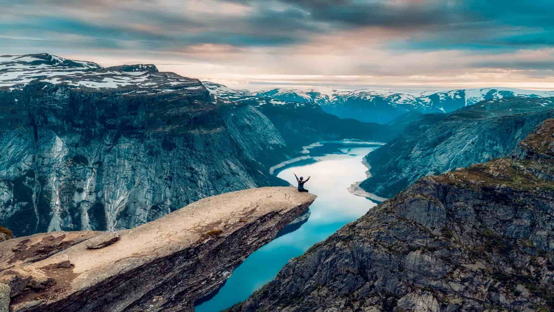 A lone hiker stands with arms raised on the iconic Trolltunga rock formation overlooking a stunning fjord and snow-capped mountains near Odda, Norway.