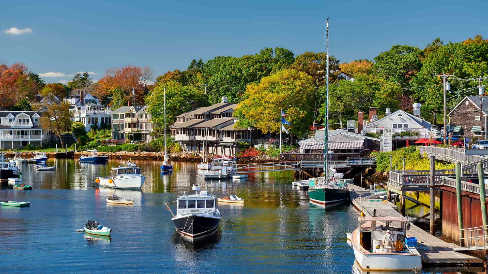 "A picturesque wide-angle shot of a serene harbor in Ogunquit, Maine, filled with boats on a sunny day with houses and autumn trees lining the shore.  "
