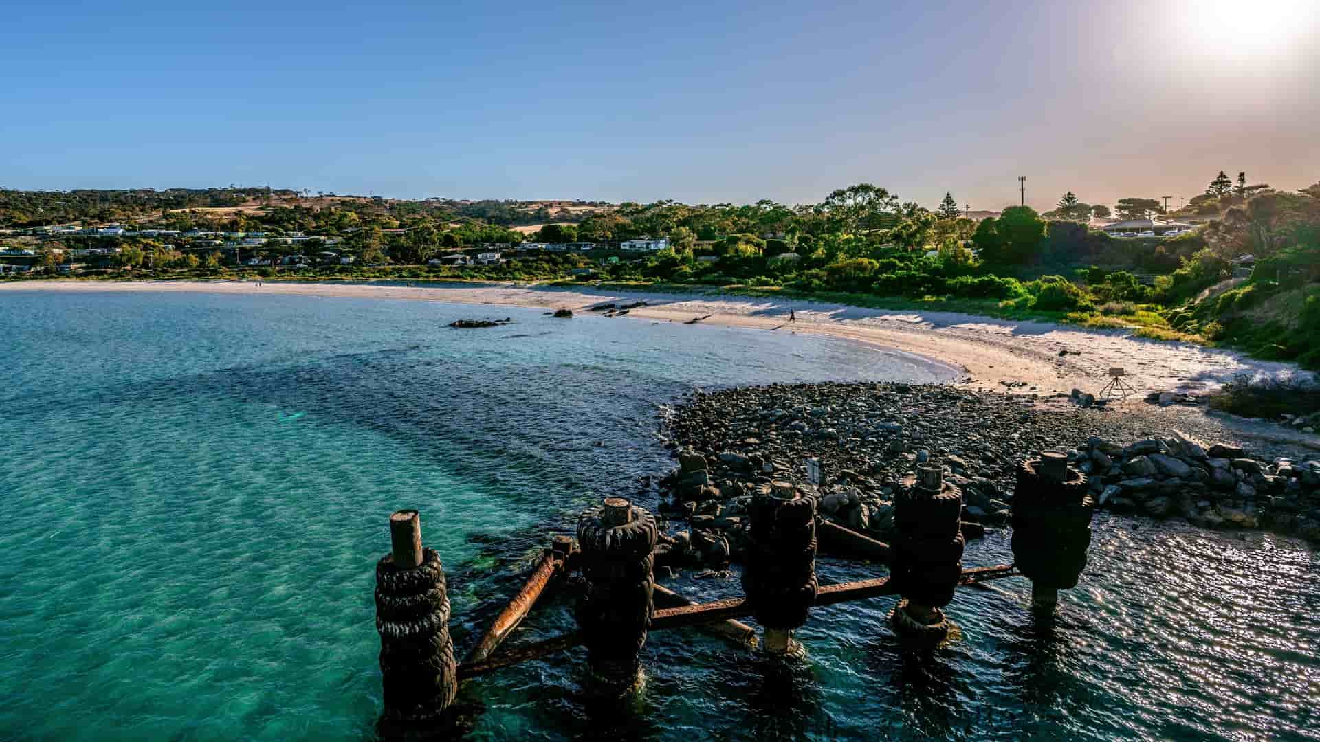 A high-angle view of the port of Penneshaw, Australia, with a pristine sandy beach, turquoise water, and rusty pier pylons in the foreground.