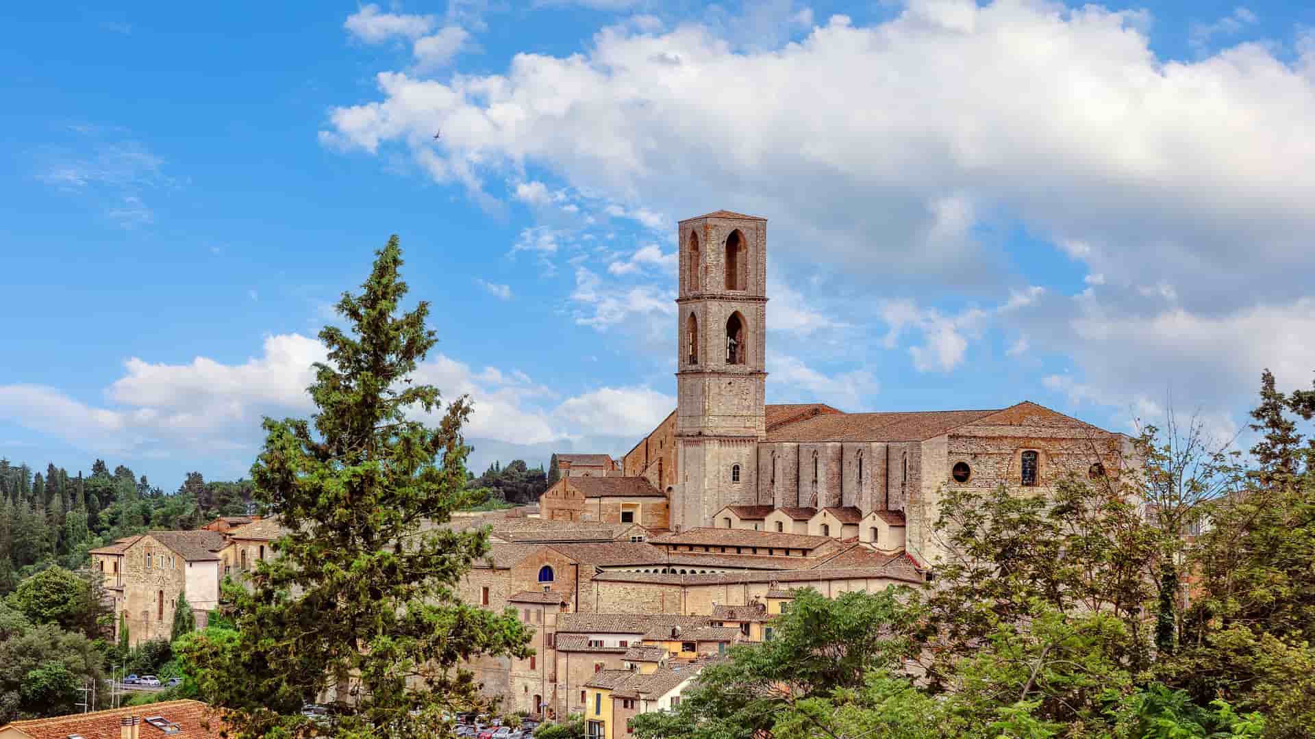 "A stunning wide-angle shot of the historic Abbey of San Pietro, a major landmark in Perugia, Italy, with its towering bell tower and medieval stone buildings surrounded by lush trees under a cloudy blue sky.  "