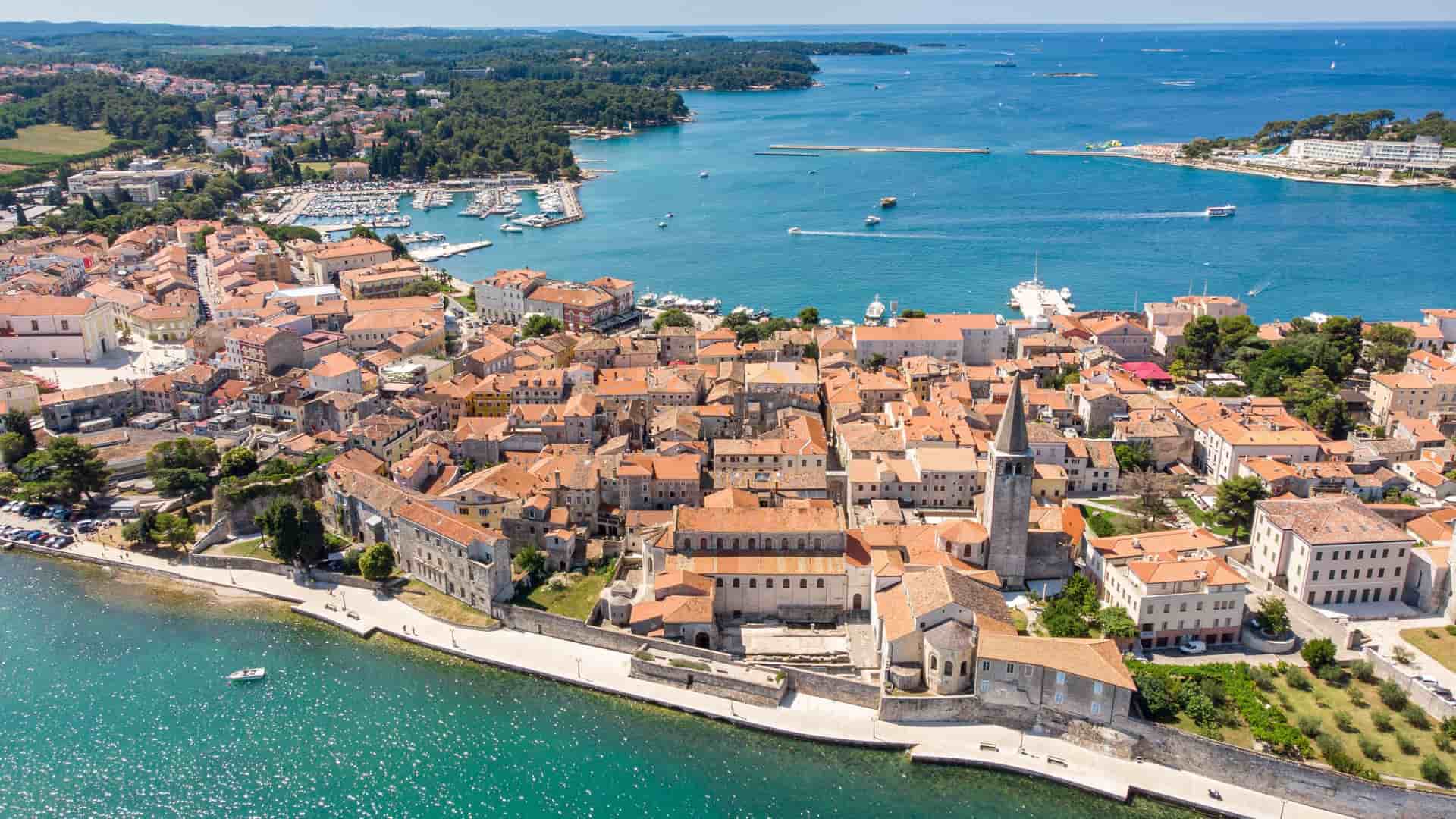 An aerial drone photograph of the historic old town of Poreč, Croatia, situated on a peninsula, showcasing its dense cluster of buildings with red-tiled roofs extending into the brilliant turquoise and blue water of the Adriatic Sea.
