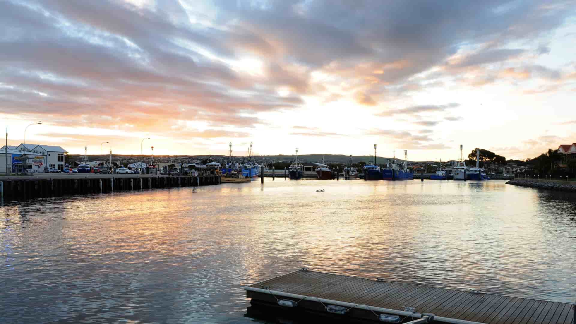 A calm harbor in Port Lincoln, South Australia, at sunrise, with a variety of fishing boats and yachts docked at the marina and the town in the distance under a dramatic, cloudy sky.