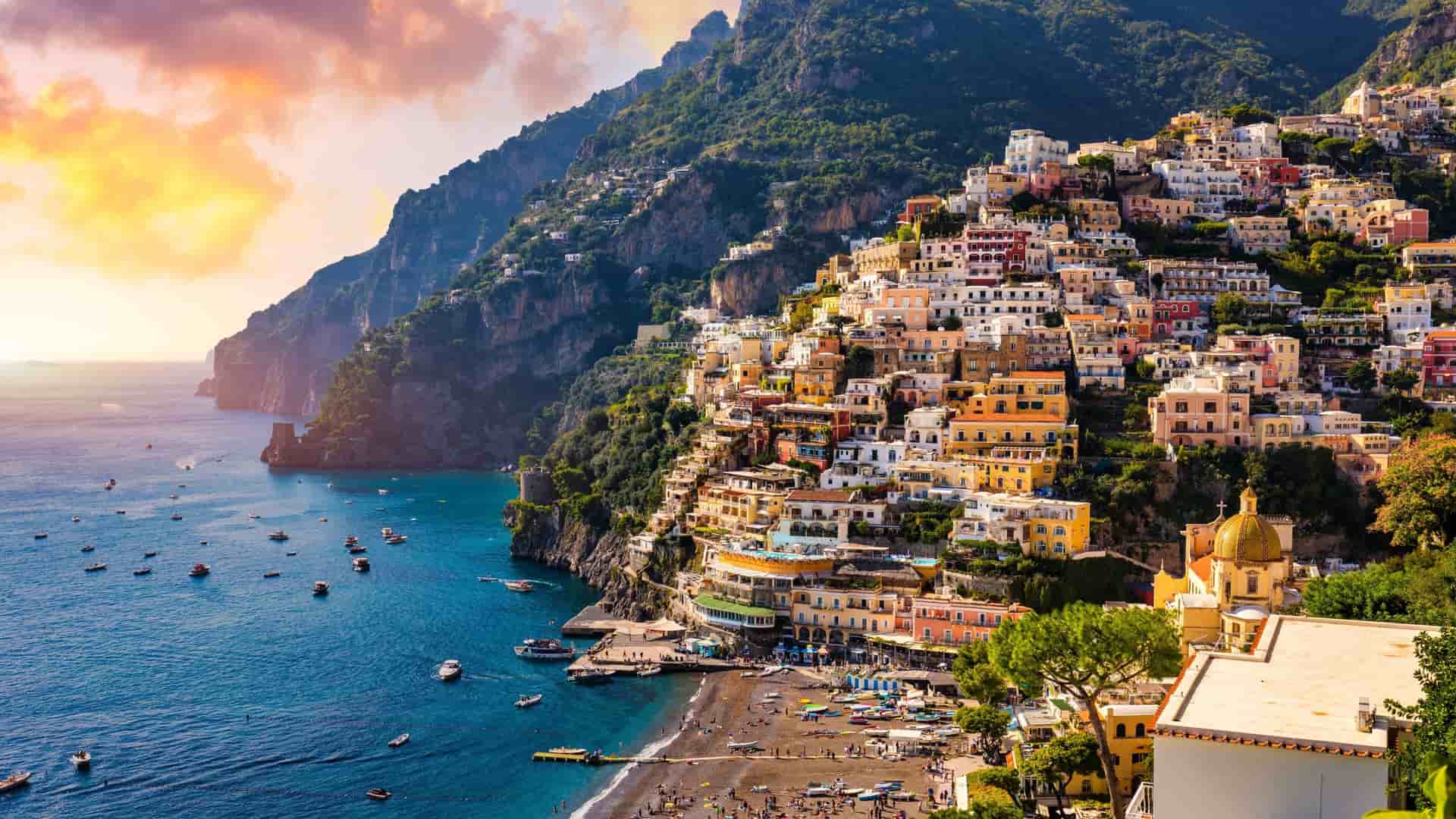 The colorful, densely packed houses of Positano cascade down a steep Amalfi Coast cliffside toward a beach and the bright blue ocean.