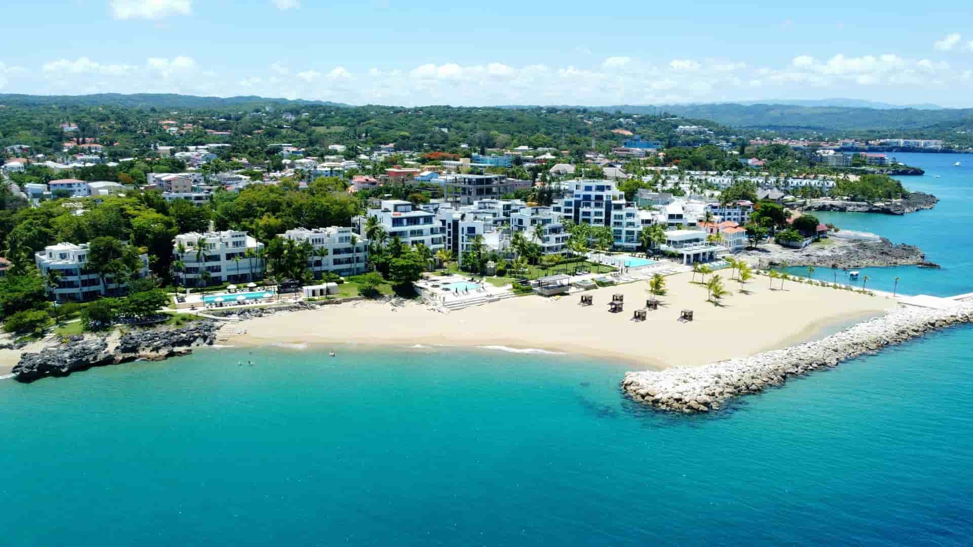 An aerial view of a luxurious resort and pristine sandy beach on the coast of Puerto Plata, Dominican Republic, with turquoise waters, lush green hills, and a cityscape in the background.