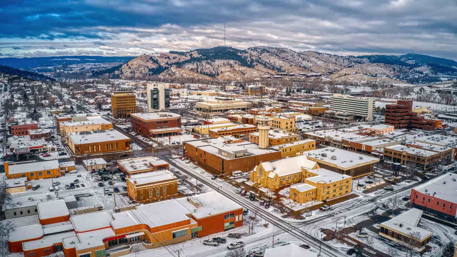 "An aerial view of downtown Rapid City, South Dakota, covered in a light blanket of snow during winter. The buildings, a mix of modern and older low-rises, have rooftops dusted with white, and the streets are visible as dark lines. The city is nestled against the backdrop of snow-covered Black Hills, with evergreen trees and patches of white snow on the slopes under a dramatic, cloudy sky.  "