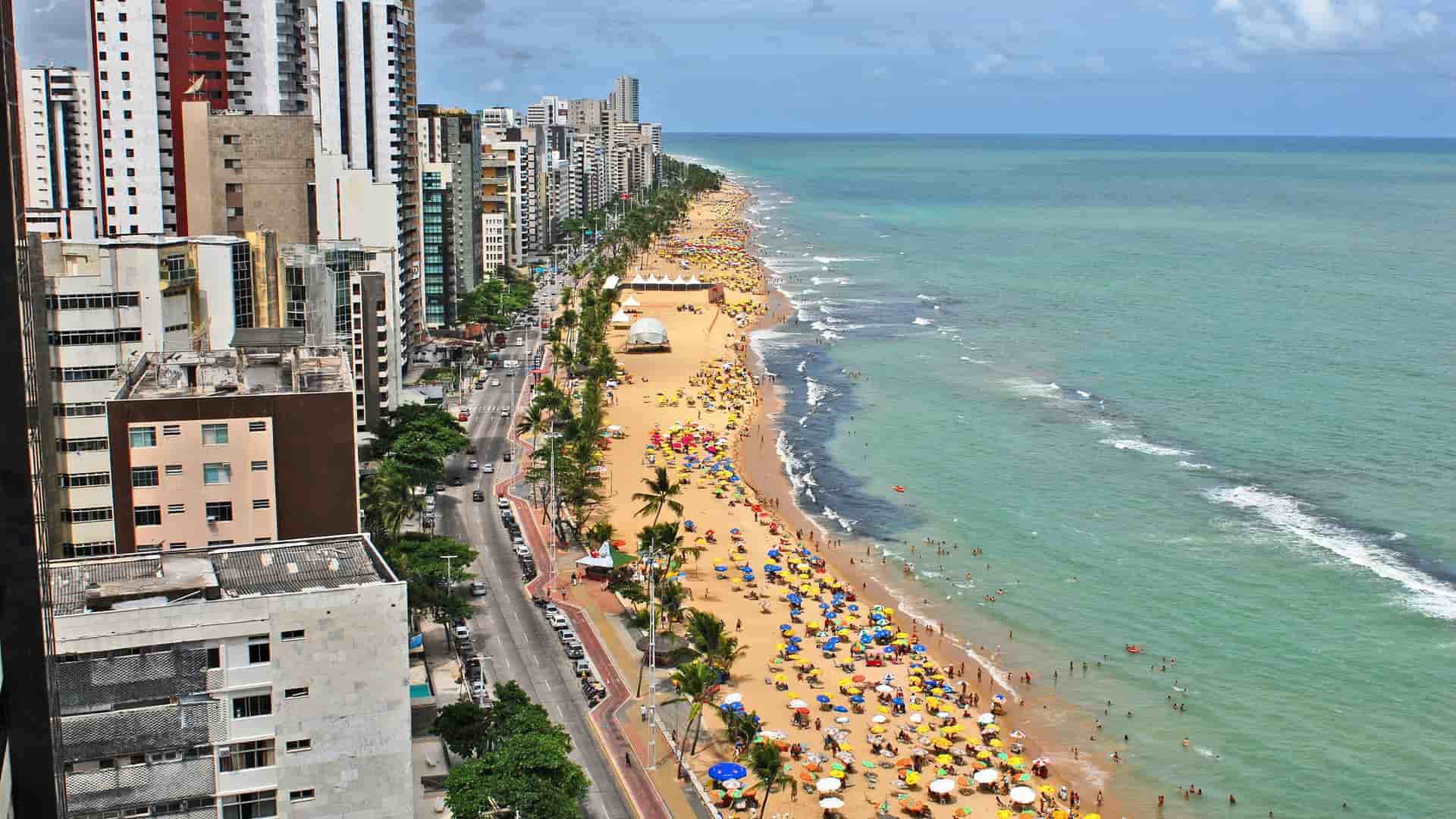An aerial view of Boa Viagem beach in Recife, Brazil, showcasing the bustling coastline with sunbathers, colorful umbrellas, and high-rise buildings along the main street.
