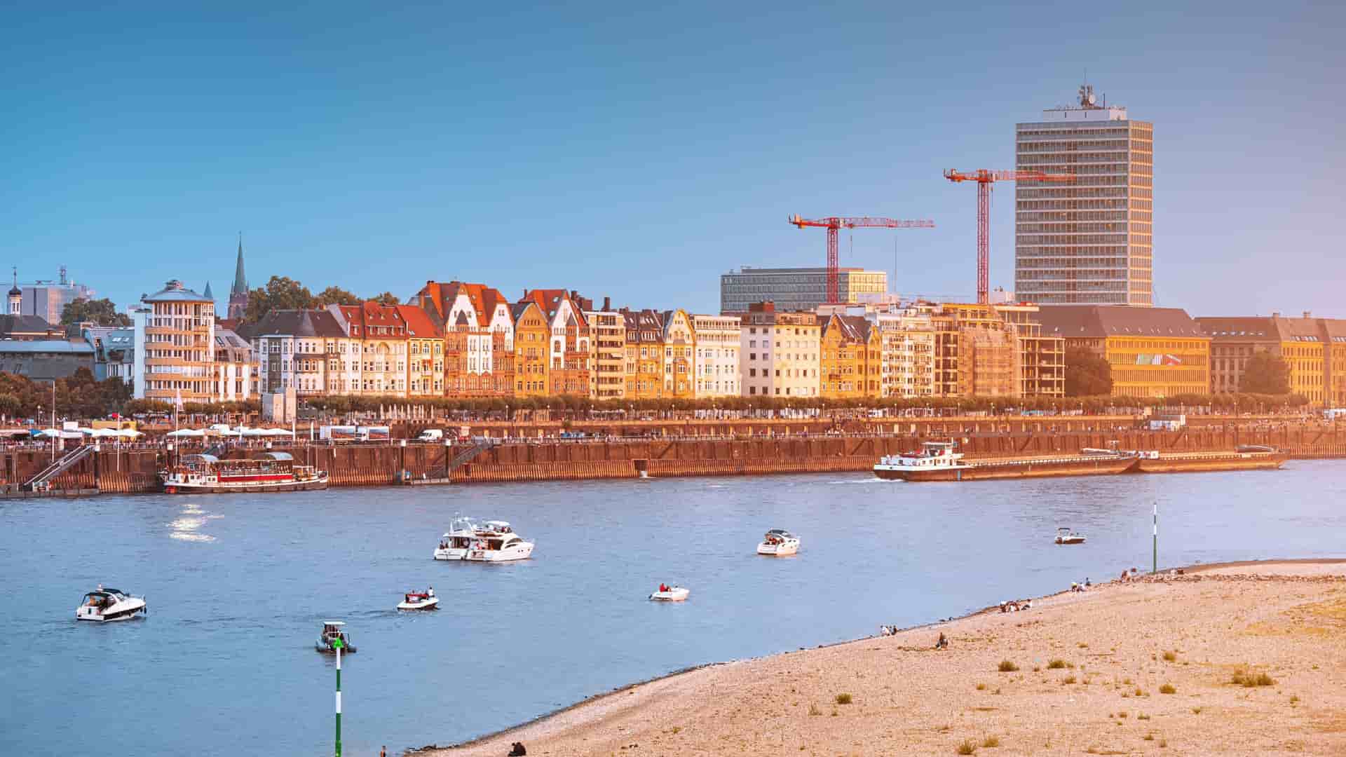 The Rhine River waterfront features boats, a sandy bank in the foreground, and historic, colorful buildings in the sunshine.
