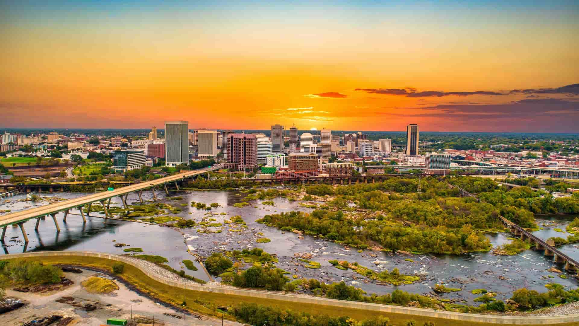 A high-angle drone view of the downtown Richmond, Virginia, USA skyline, showcasing a cluster of modern high-rise buildings and older brick structures. The cityscape is situated along the banks of the James River, with a bridge or railroad trestle visible crossing the water in the foreground or midground. The image is taken during the day, likely featuring a clear or partly cloudy sky.