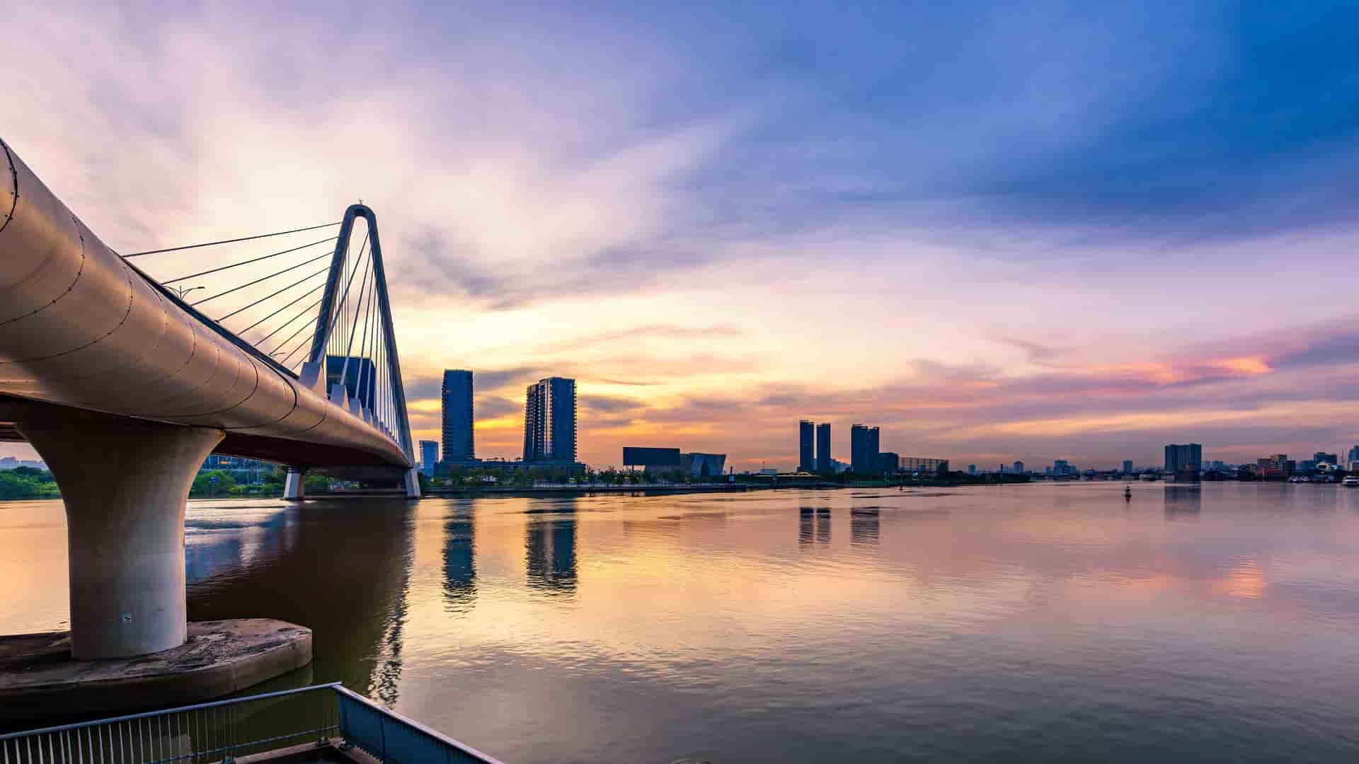 A scenic sunset over the Saigon River, with a modern suspension bridge in the foreground and the silhouette of the Ho Chi Minh City skyline in the background.