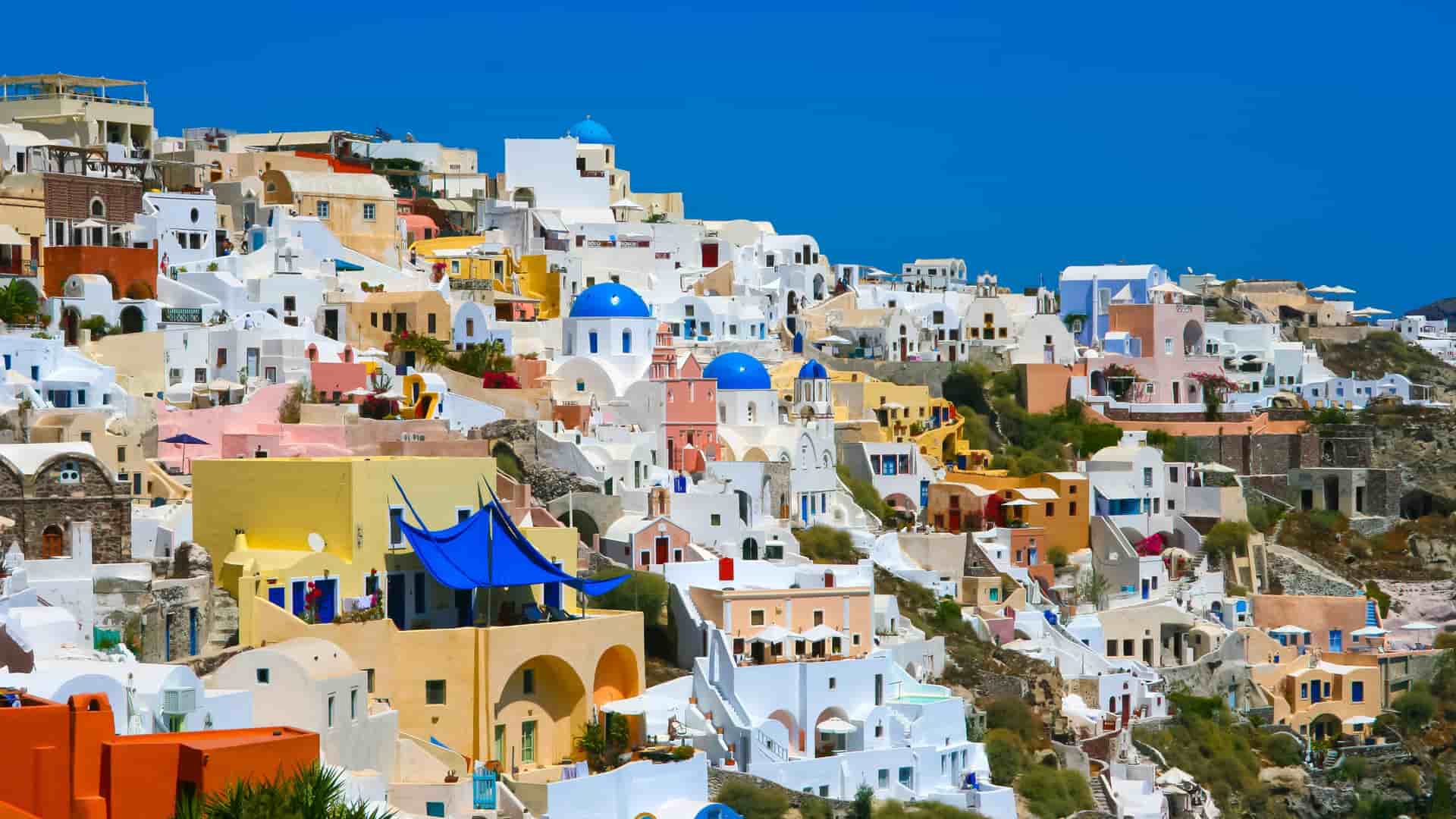 A stunning photograph of the iconic cliffside village of Oia in Santorini, Greece, with its whitewashed buildings and blue-domed churches cascading down the volcanic caldera, set against a deep blue sky.