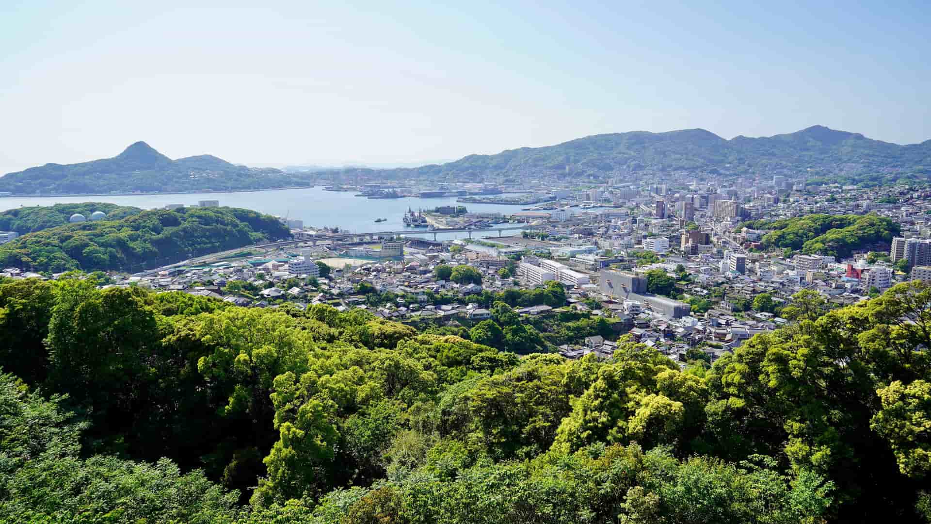 A scenic high-angle view of the port city of Sasebo in Nagasaki, Japan, nestled between lush green forests and the serene blue bay.
