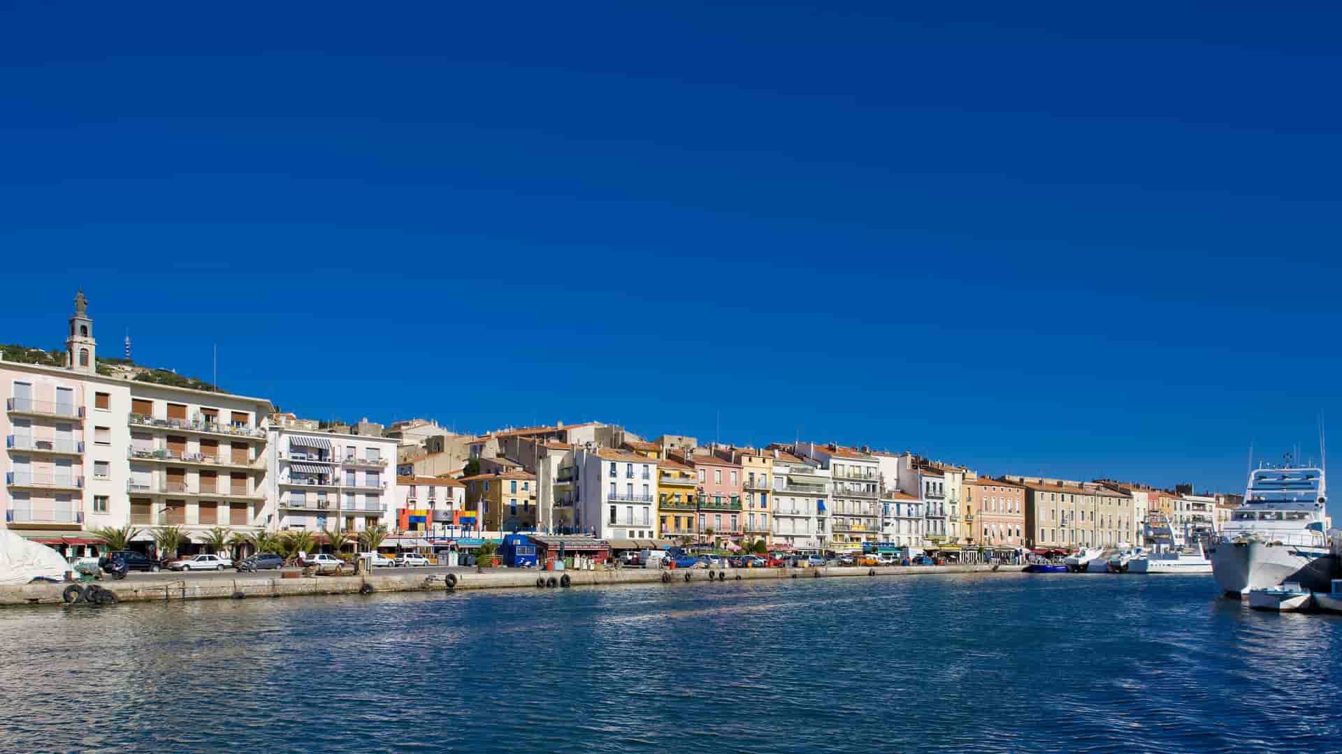 A vibrant daytime view of the Sète waterfront in France, featuring colorful buildings along a canal with boats moored in the harbor under a clear blue sky.