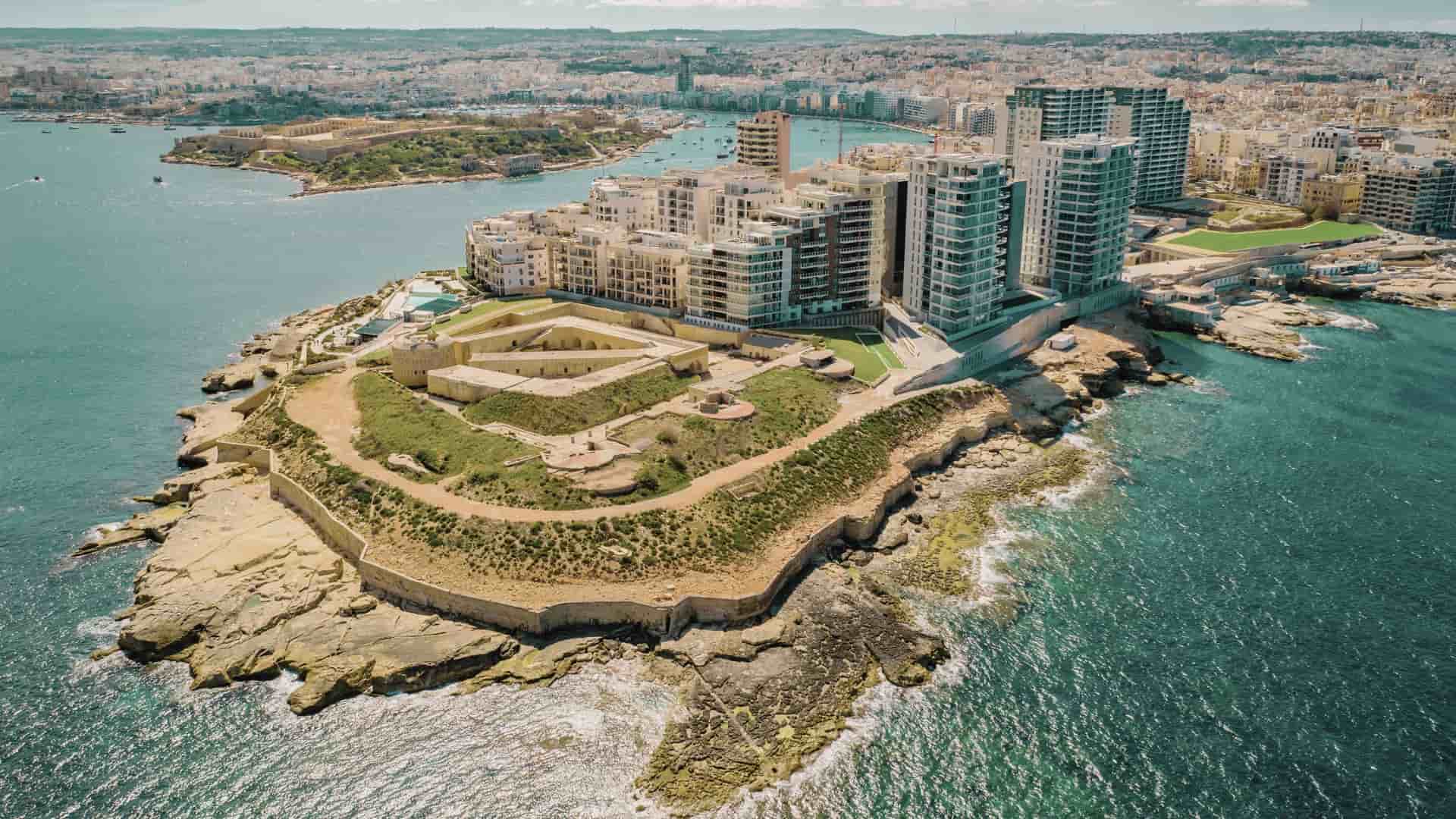 An aerial view of the densely built coastal city of Sliema, Malta, focusing on a concentration of limestone buildings with traditional Maltese enclosed balconies and modern, multi-story developments. The clear, blue waters of the Mediterranean Sea or the Marsamxett Harbour would be visible in the background or along the waterfront.
