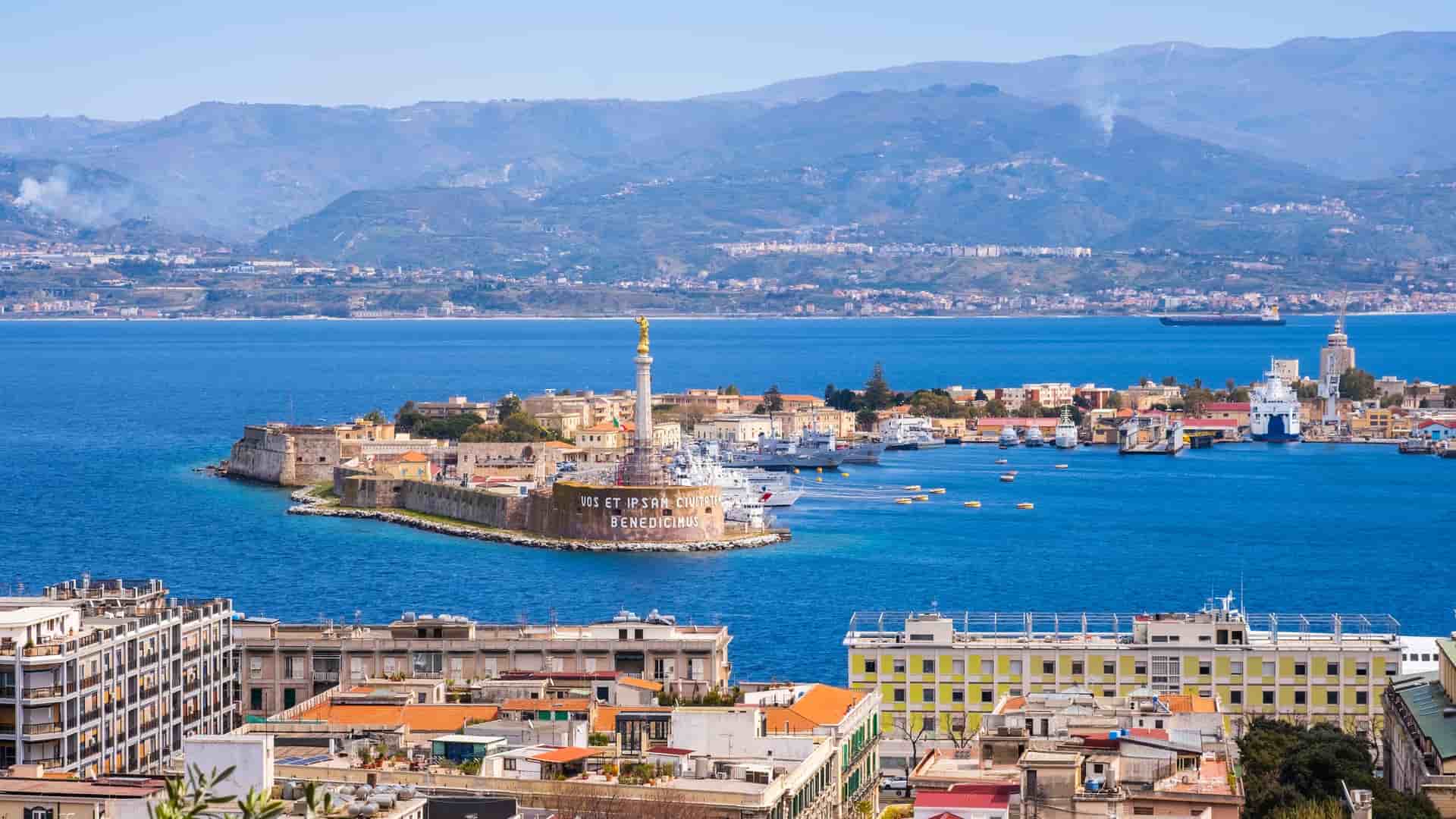 A wide-angle view of the bustling harbor of Messina in Sicily, Italy, with the iconic gold statue of the Madonna della Lettera on a monument overlooking the Strait of Messina.