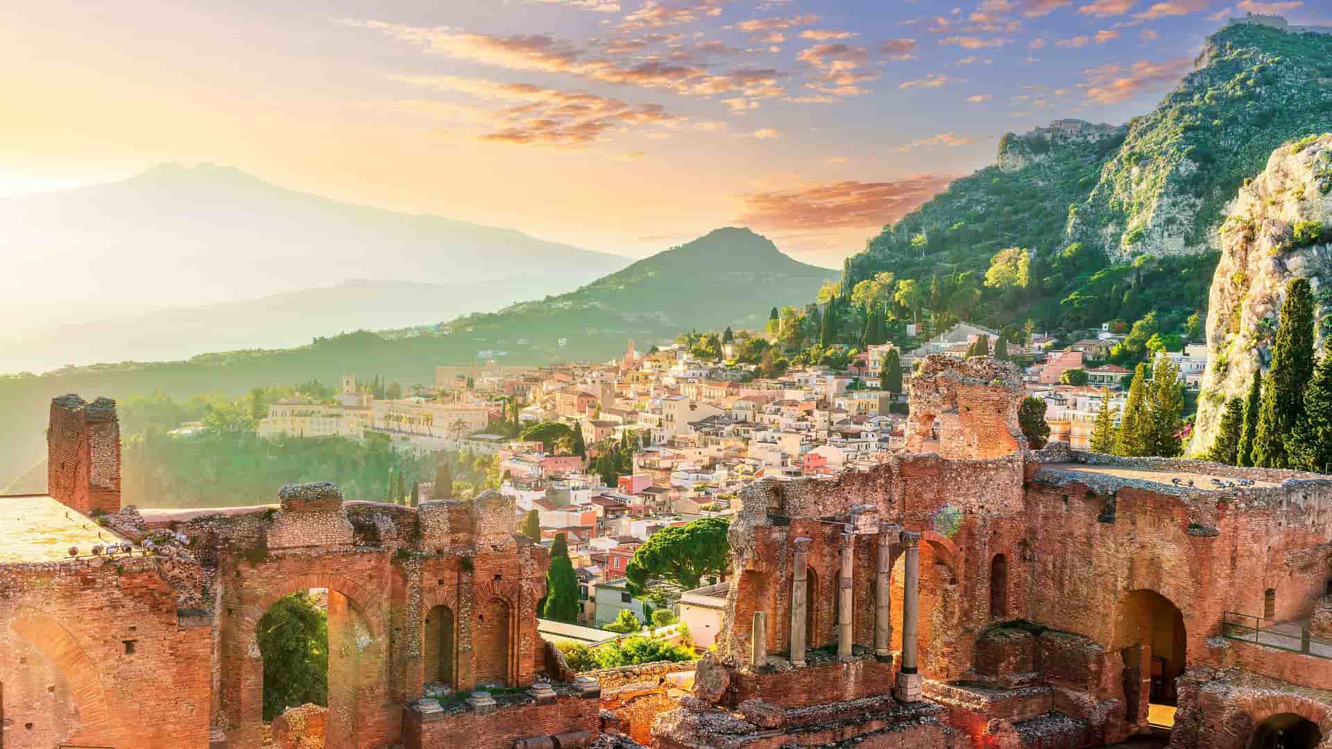 A panoramic sunset view from the ancient Greek Theatre of Taormina in Sicily, Italy, overlooking the city of Giardini Naxos, with Mount Etna in the background under a beautiful orange and pink sky.