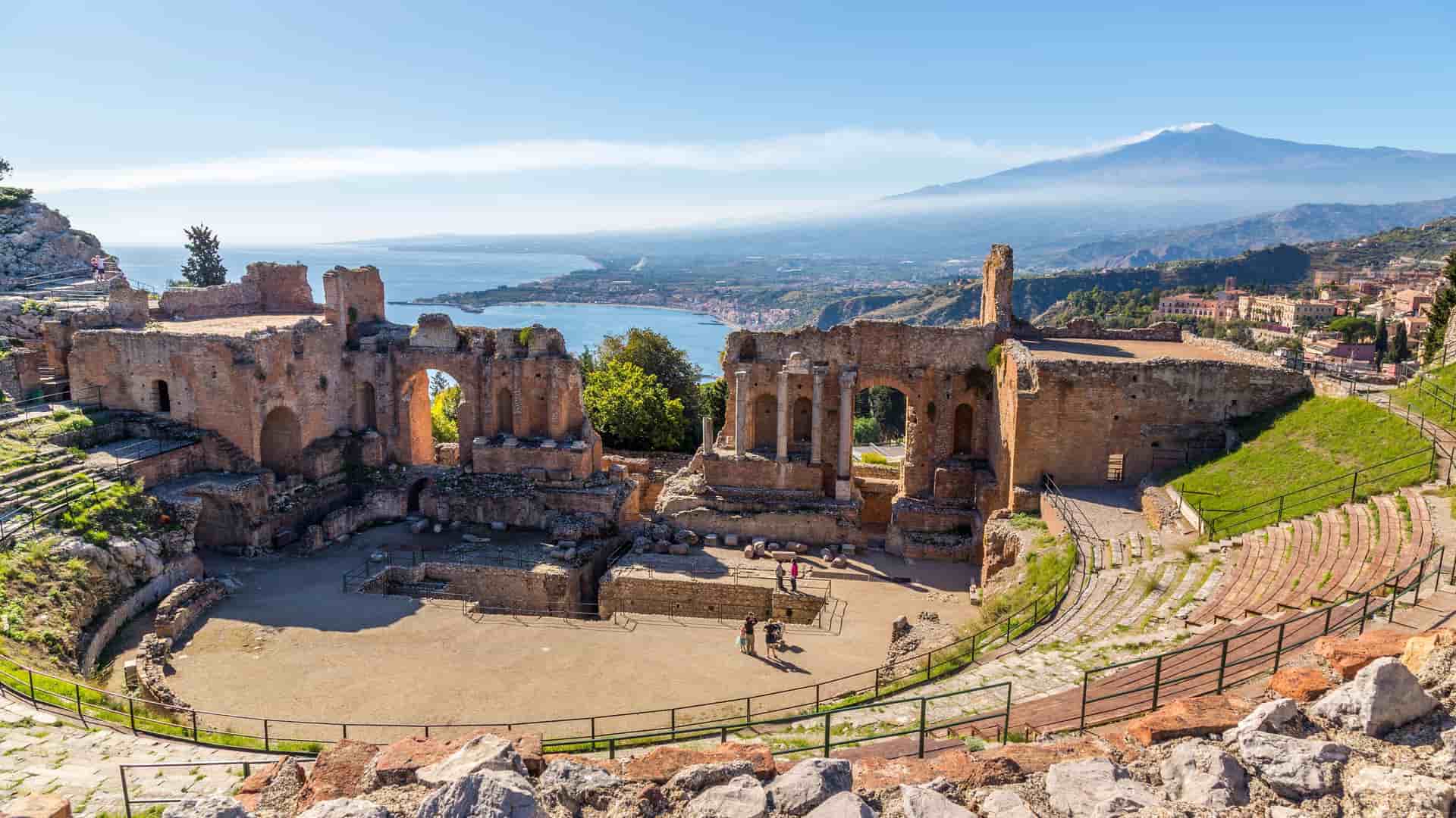 A scenic view of the ancient Greek theater ruins in Taormina, Sicily, with a panoramic backdrop of the bay, coastline, and majestic Mount Etna.