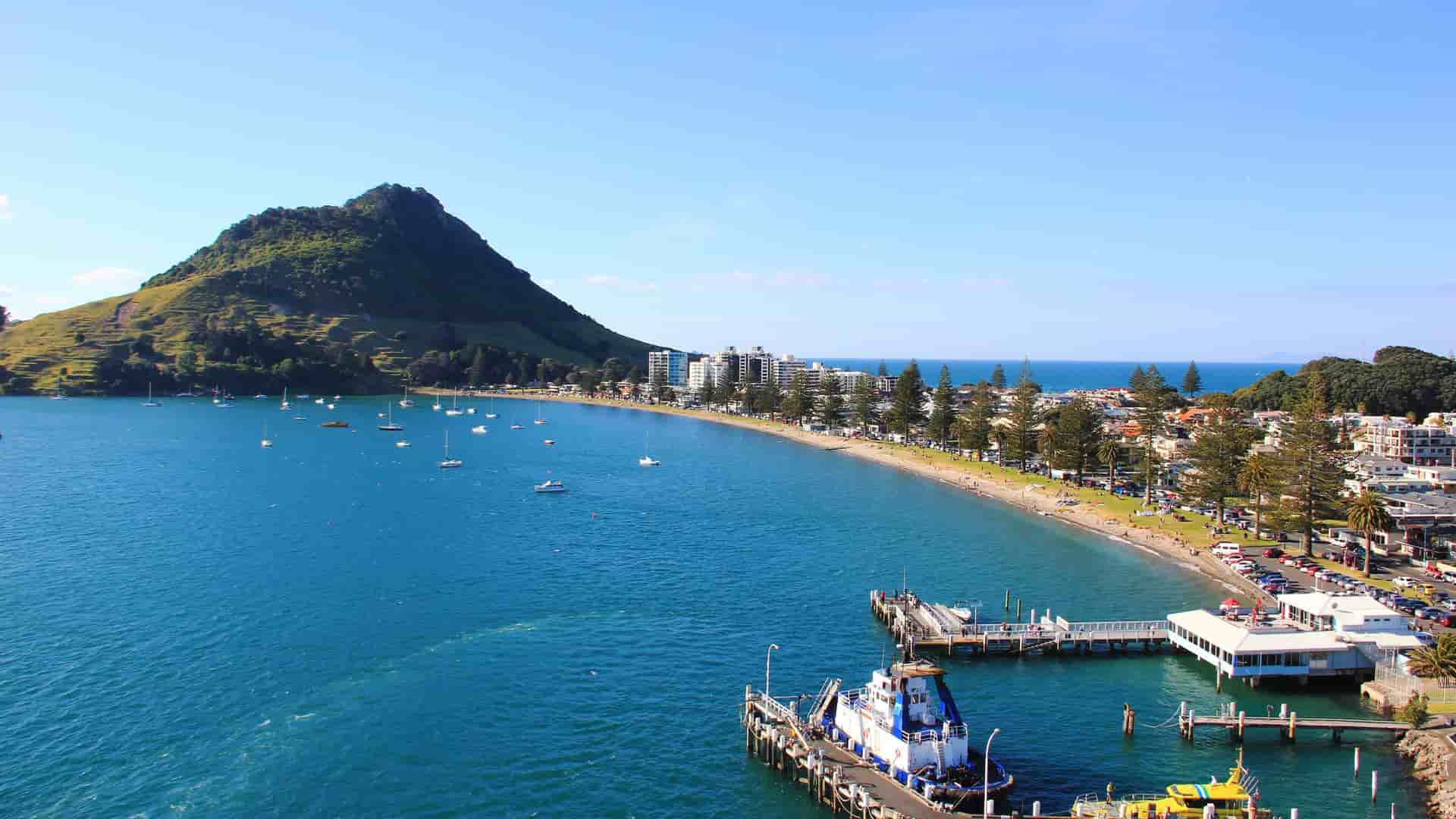 A panoramic view of the beach and harbor in Tauranga, New Zealand, with Mount Maunganui in the background and boats anchored in the clear blue water.