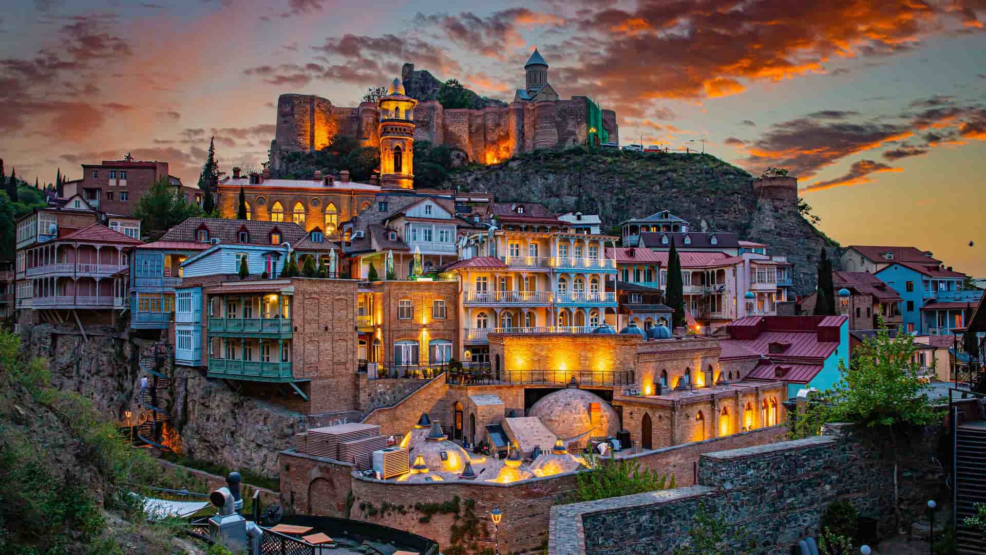 "A beautiful twilight shot of the old town of Tbilisi, Georgia, with the historic Narikala Fortress and a church perched on a cliff above the city, and the traditional brick buildings glowing with light.  "