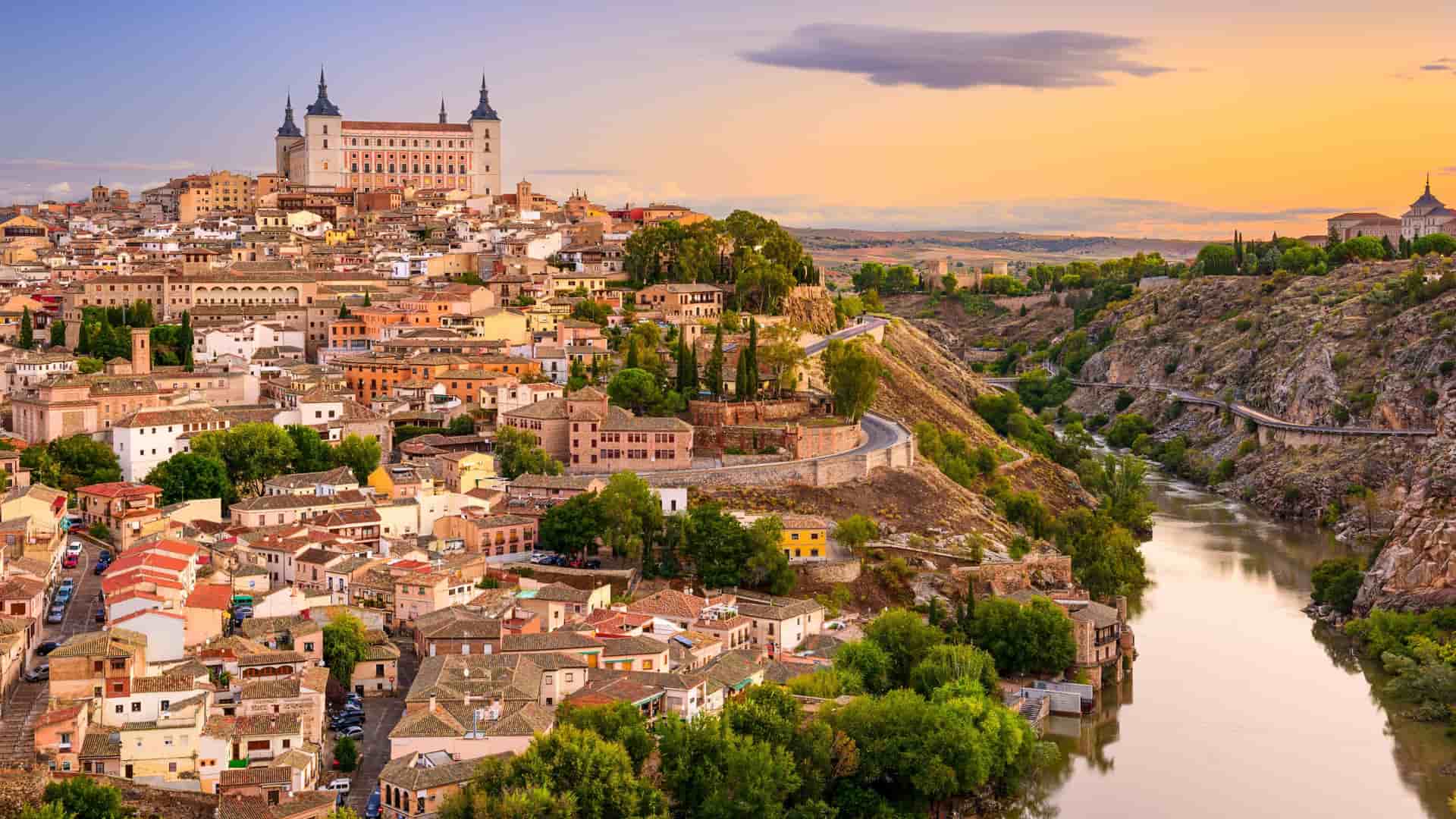 A panoramic view of the medieval city of Toledo, Spain, with the historic Alcántara Bridge spanning the Tagus River and the Alcázar of Toledo fortress rising on a hill.