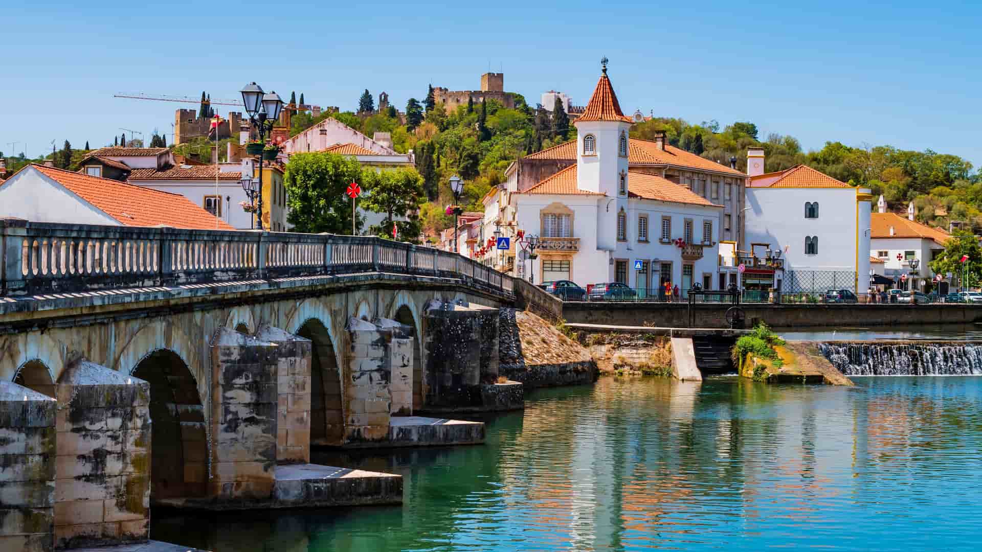 A picturesque view of the medieval city of Tomar, Portugal, with a stone bridge spanning the Nabão River, historic white buildings with orange-tiled roofs, and the Convento de Cristo castle on the hill.