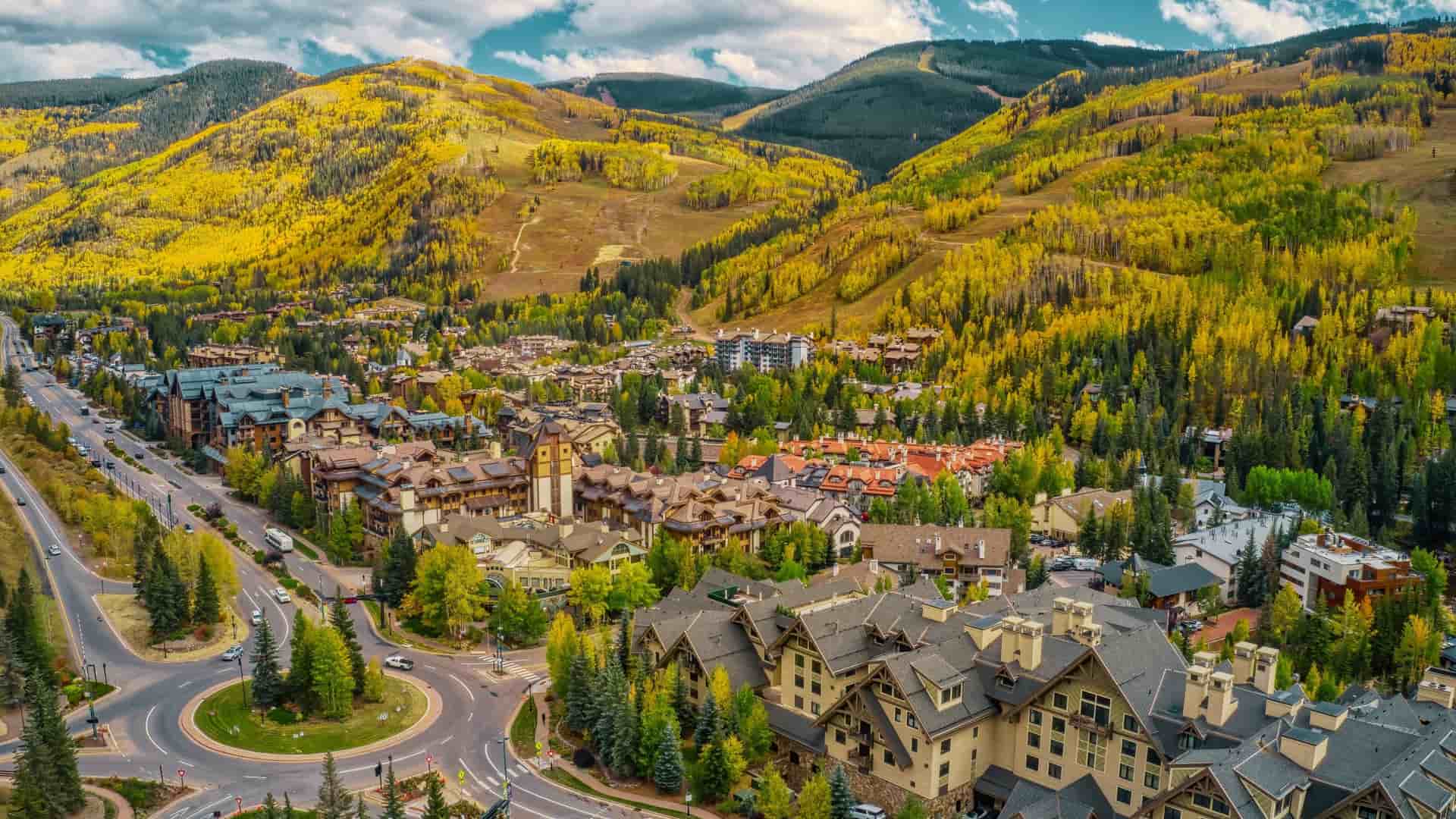 An aerial view of Vail, Colorado during the autumn season. The image captures the valley floor where the town is situated, with the Gore Creek winding through it and the distinctive, European-style village architecture visible in the center. The surrounding mountainsides are ablaze with fall foliage, primarily vibrant golden-yellow Aspen trees contrasted against the dark green of pine and spruce evergreens, all under a clear sky.
