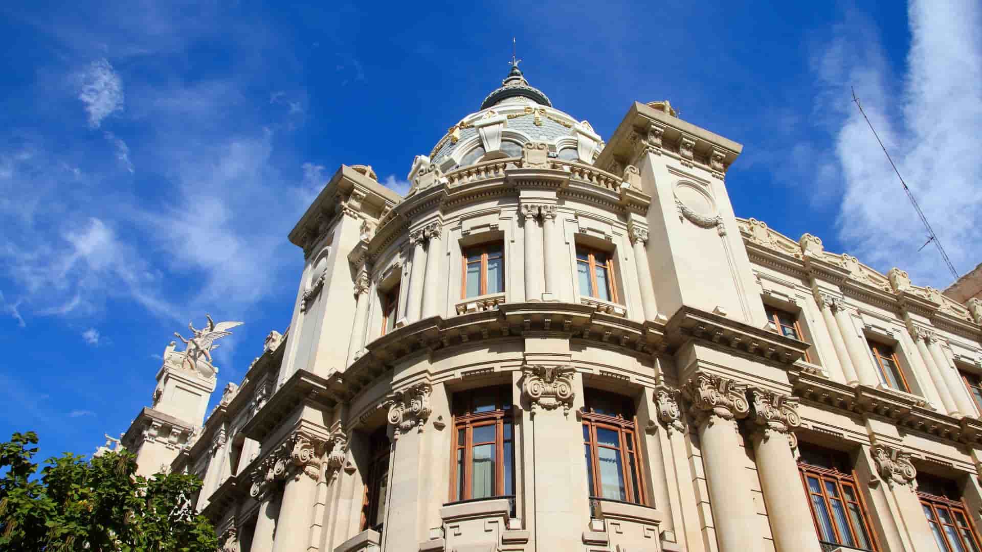 A striking low-angle shot of a classic Art Nouveau building, the Estación del Norte train station in Valencia, Spain, featuring intricate carvings and a blue-tiled dome against a bright blue sky.