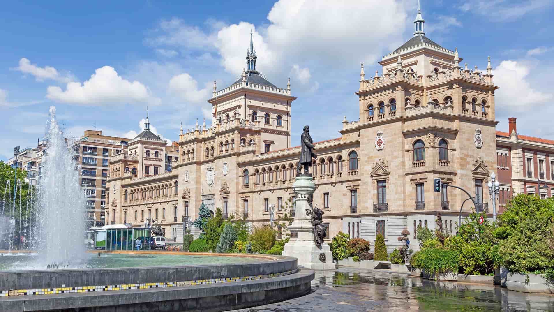 "A sunny daytime shot of the Royal Palace of Valladolid, Spain, with a large fountain in the foreground and a bronze statue in front of the ornate, historic brick building.  "