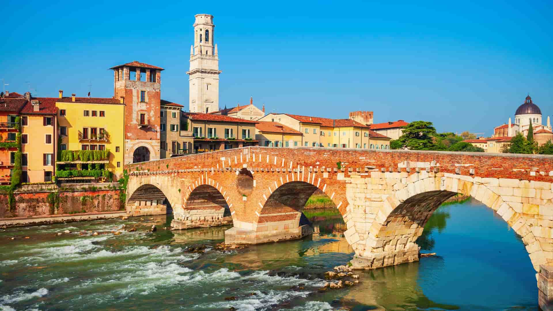 A sunny view of the historic Ponte Pietra bridge over the Adige River in Verona, Italy, with the medieval city buildings and the bell tower of the Verona Cathedral in the background.