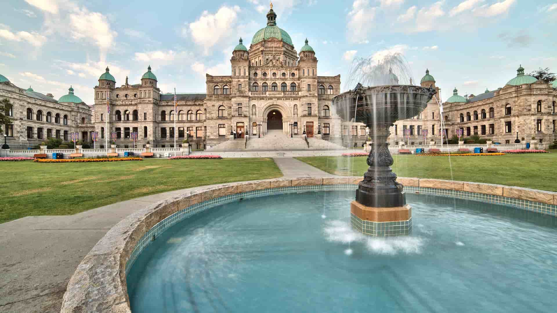 A grand view of the British Columbia Parliament Buildings in Victoria, with a large, ornate fountain in the foreground and a well-manicured lawn under a bright, cloudy sky.