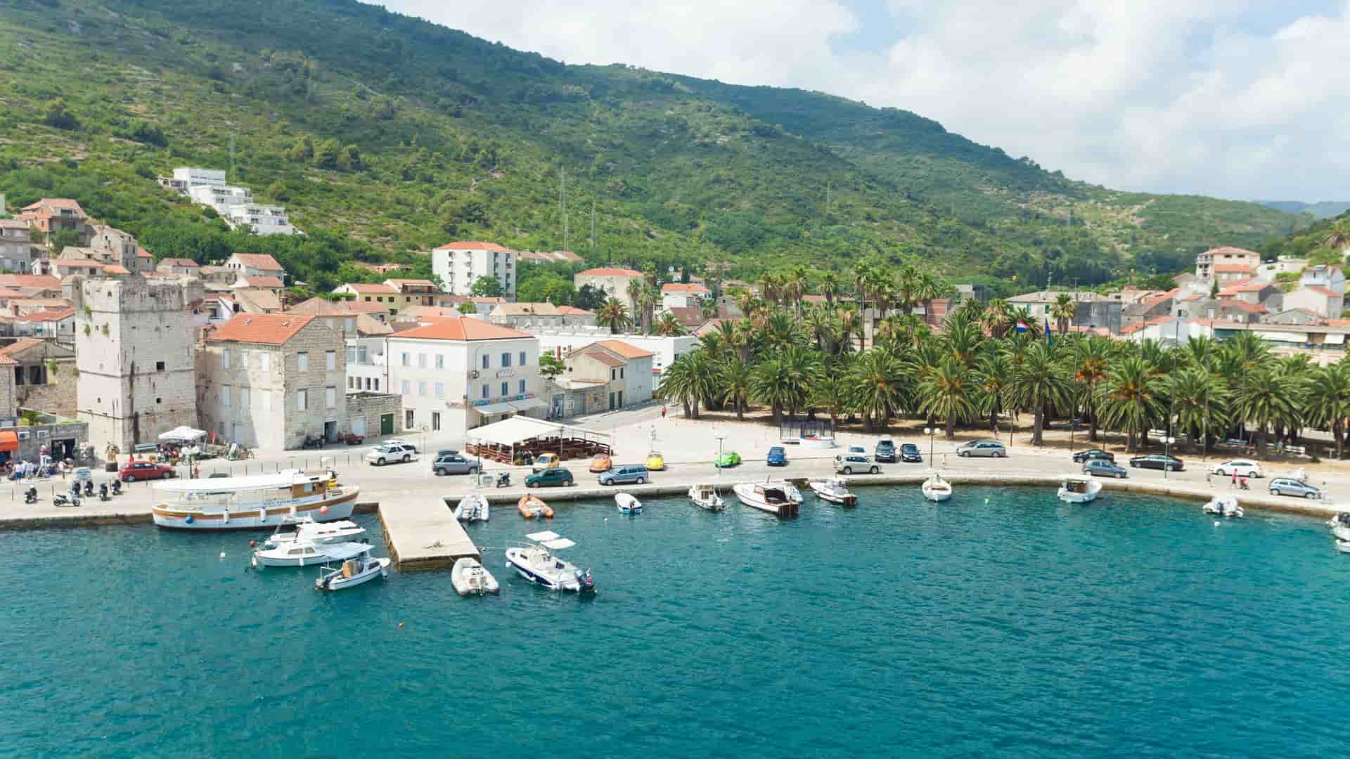 A scenic view of Vis harbor in Croatia, with small boats docked at the pier, surrounded by historic buildings and a lush green hillside.