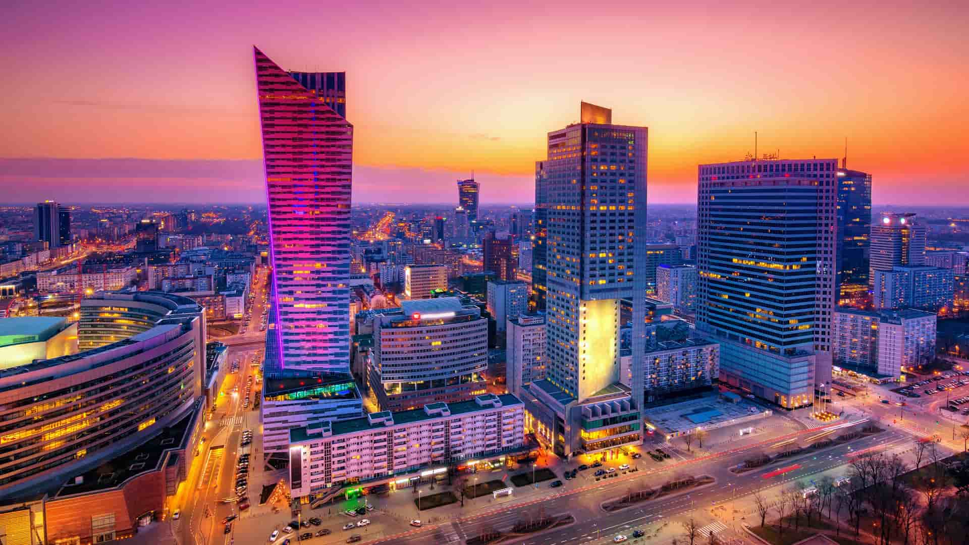 "A stunning panoramic shot of the modern city skyline of Warsaw, Poland, at dusk, with the high-rise buildings illuminated against a vibrant orange and purple sky.  "