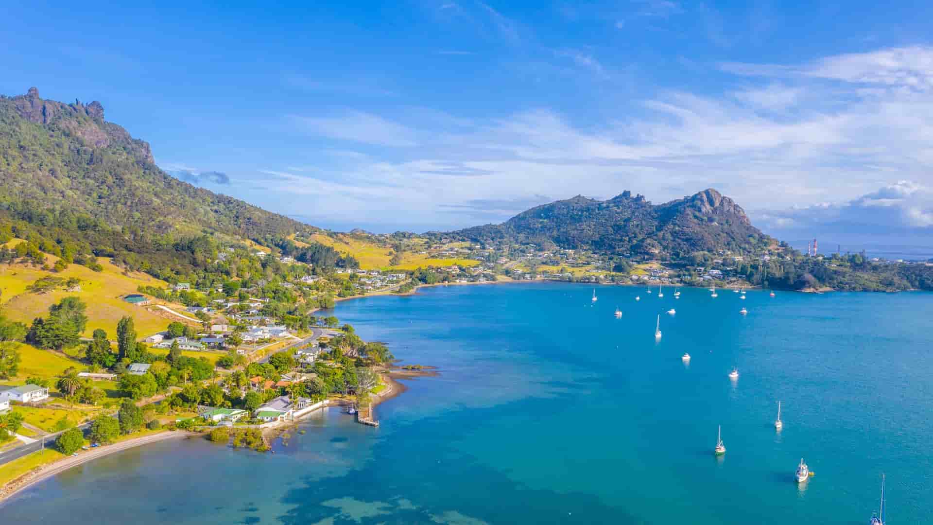 An aerial view of the turquoise blue Whangarei Harbour in New Zealand. The surrounding coastline has small communities and is backed by green, rolling hills and a distinctive rocky peak.