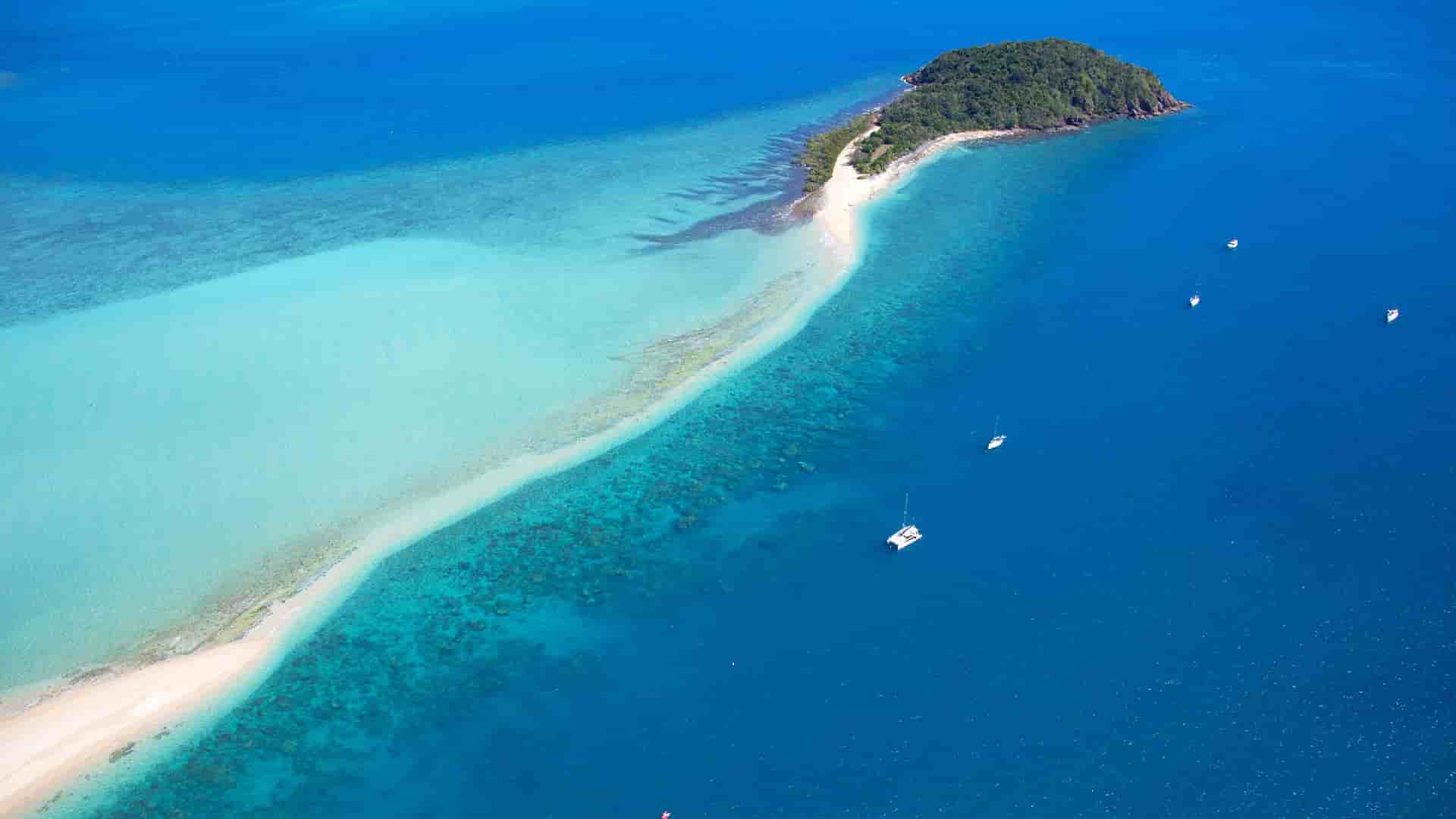 An aerial view of a white sand beach and a small island in the turquoise waters of the Whitsunday Islands, Australia, with a catamaran and other sailboats offshore.