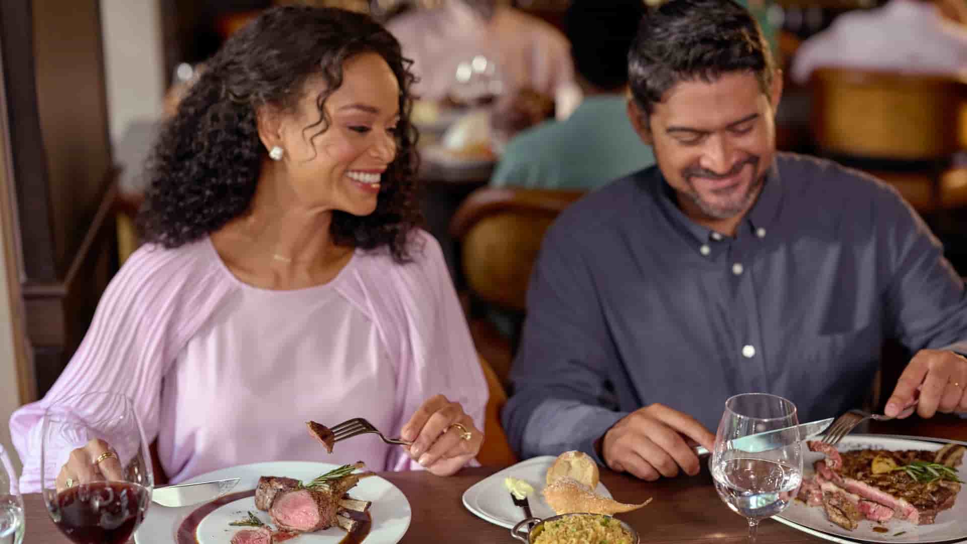 A couple enjoying a meal together on the Royal Caribbean International Symphony of the Seas ship, with the woman smiling at the man as she holds a fork over a rack of lamb and the man looks down at his plate of steak.